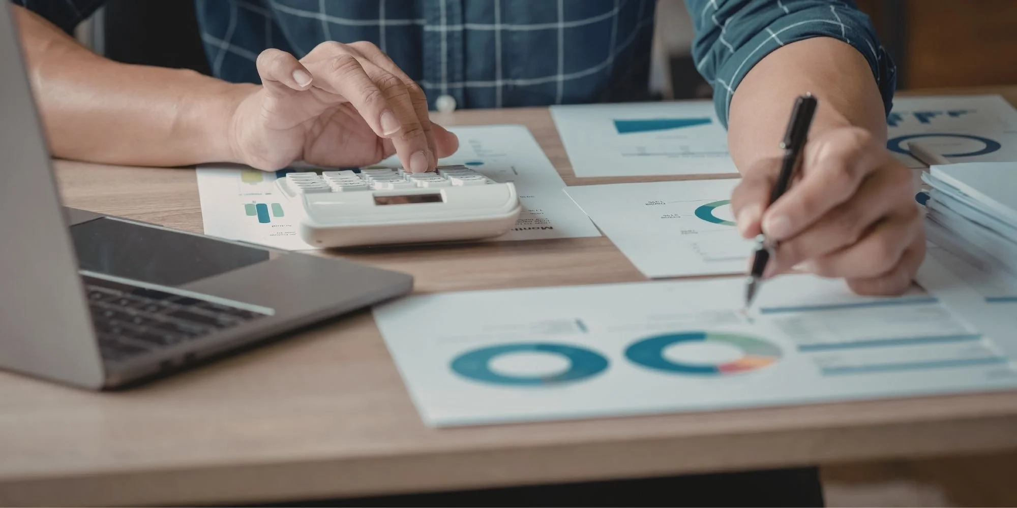 Person working at a desk with financial documents, using a calculator and writing on papers with graphs and charts.