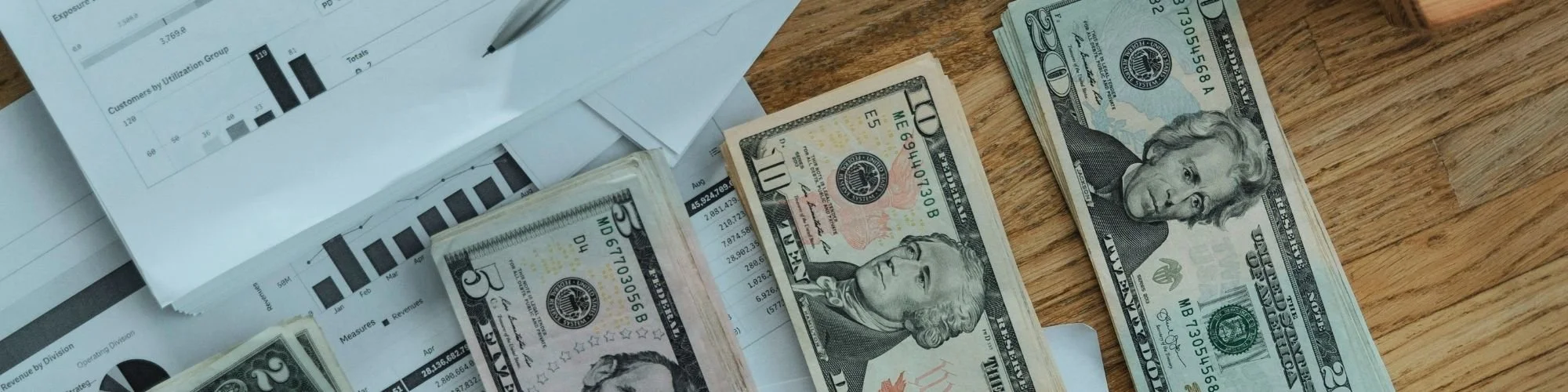 Stacks of US dollar bills ($20, $10, and $5) on a wooden desk, with financial papers and a pen.