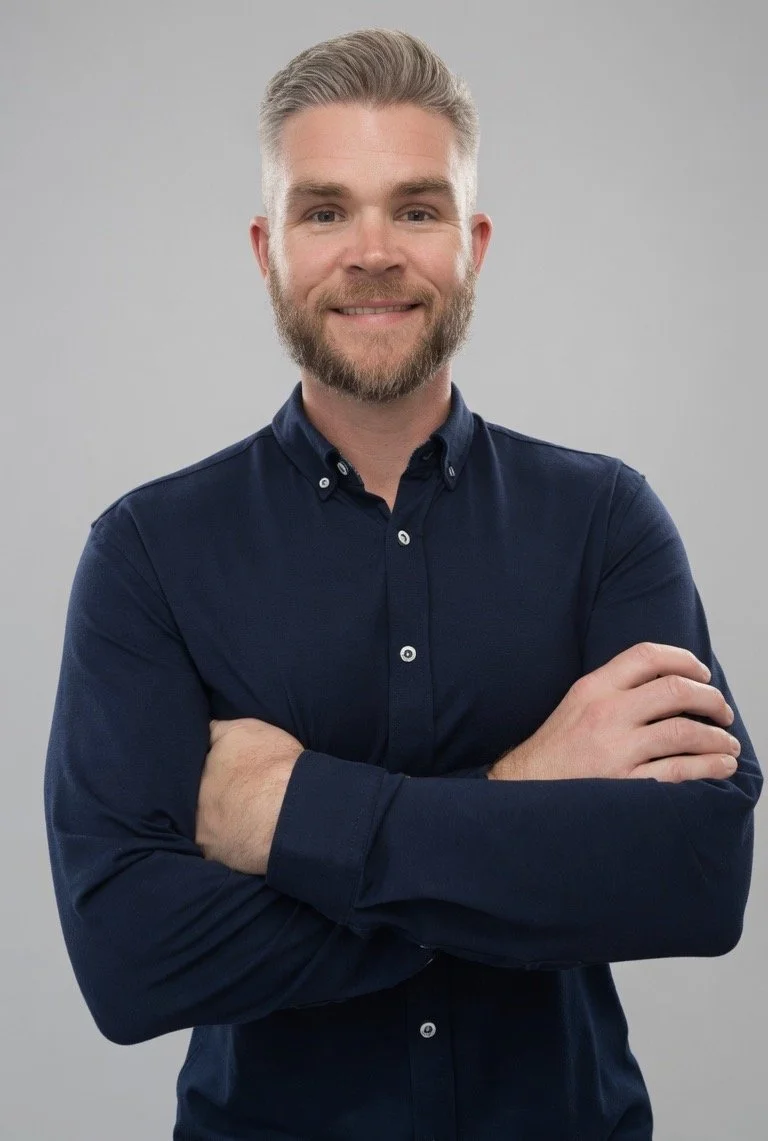 A man with short, light gray hair and a beard, smiling, wearing a dark blue button-up shirt, standing against a plain gray background.