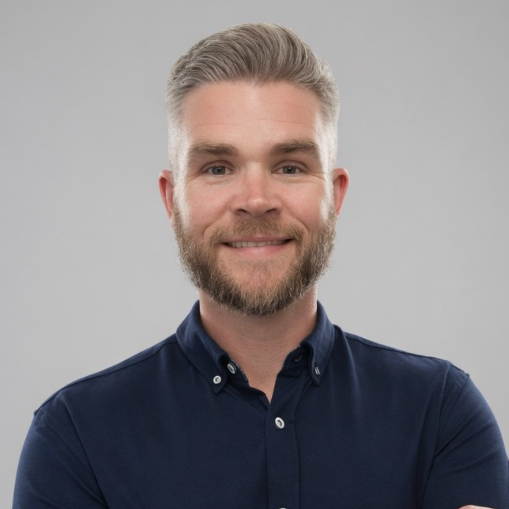 Headshot of a smiling man with short, light-colored hair and a beard, wearing a navy blue button-up shirt, against a plain gray background.