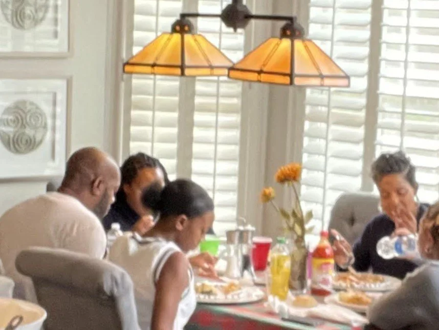 Family of four sits around a dining table with bright sunlight filtering in through windows in the background