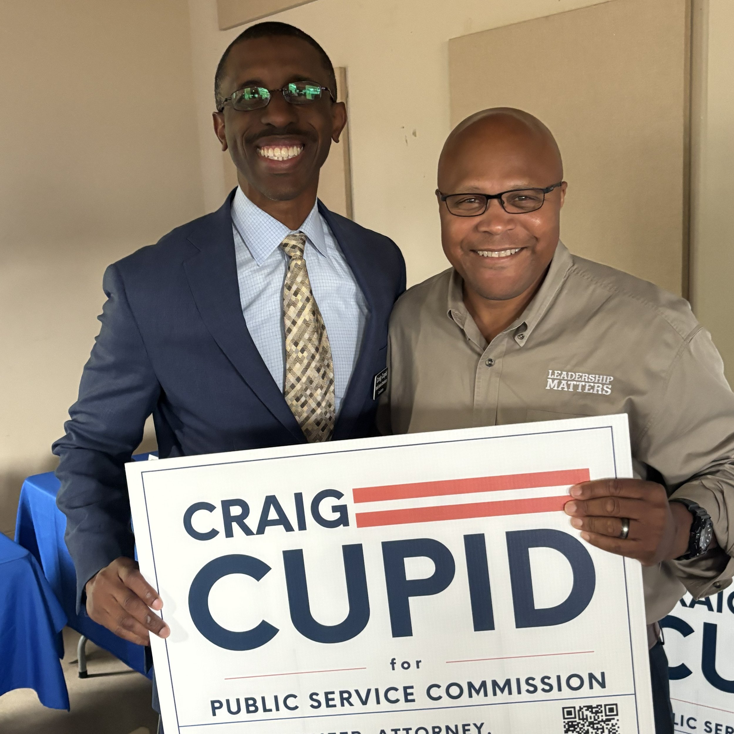 Two men smiling, one in a suit and tie and glasses, the other in a beige shirt and glasses, holding a political campaign sign that reads 'Craig for Public Service Commission' with a QR code at the bottom.