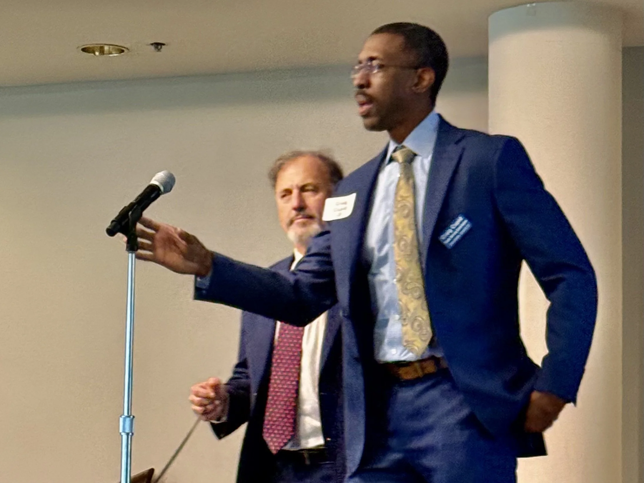 tall Black man in a sharp navy suit with yellow tie steps up to take the microphone
