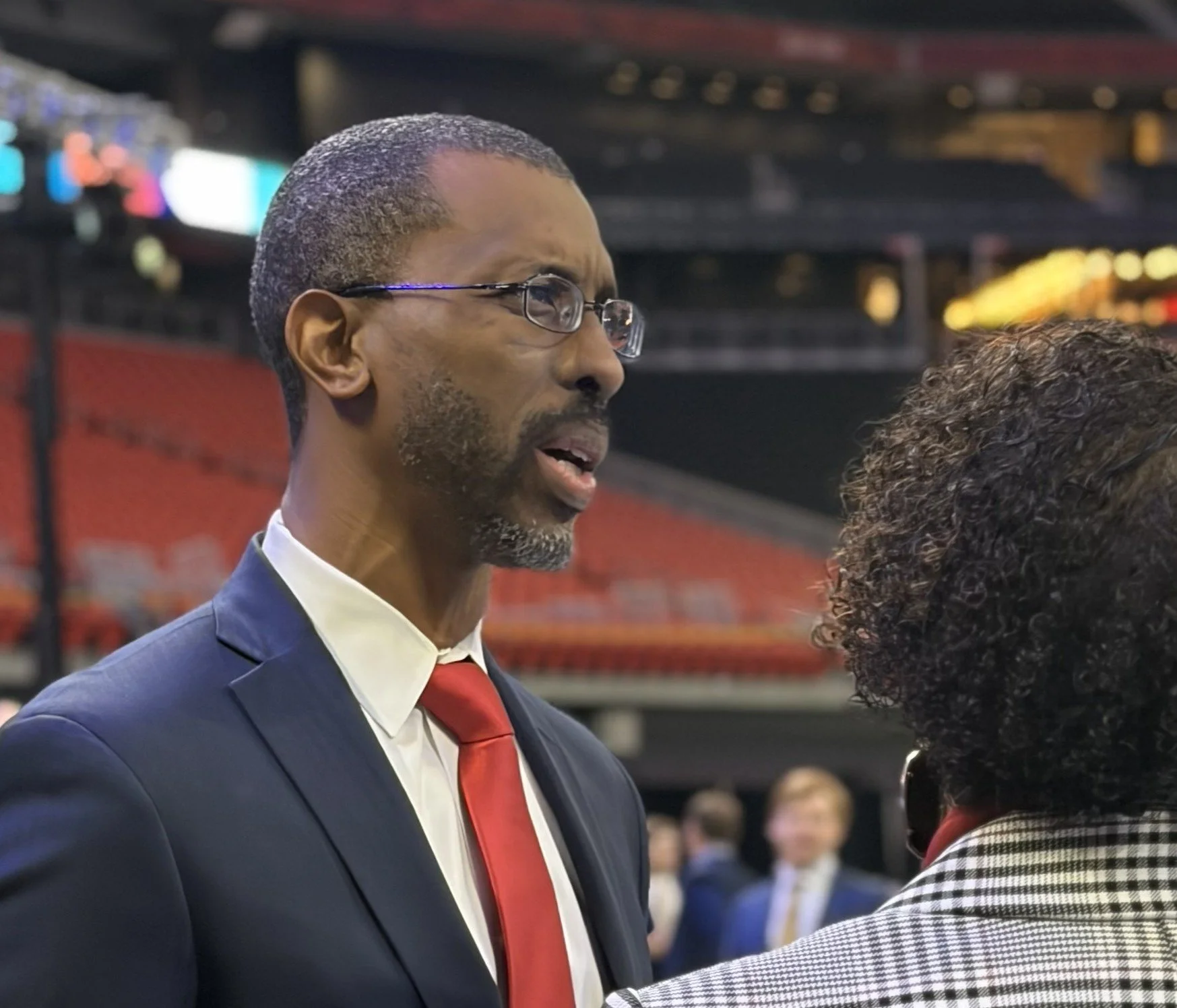 Black man in a navy suit and red tie with glasses talks to a woman in a stadium
