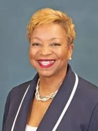 Professional woman with short blonde hair, smiling, wearing a dark blazer with white trim and a pearl necklace