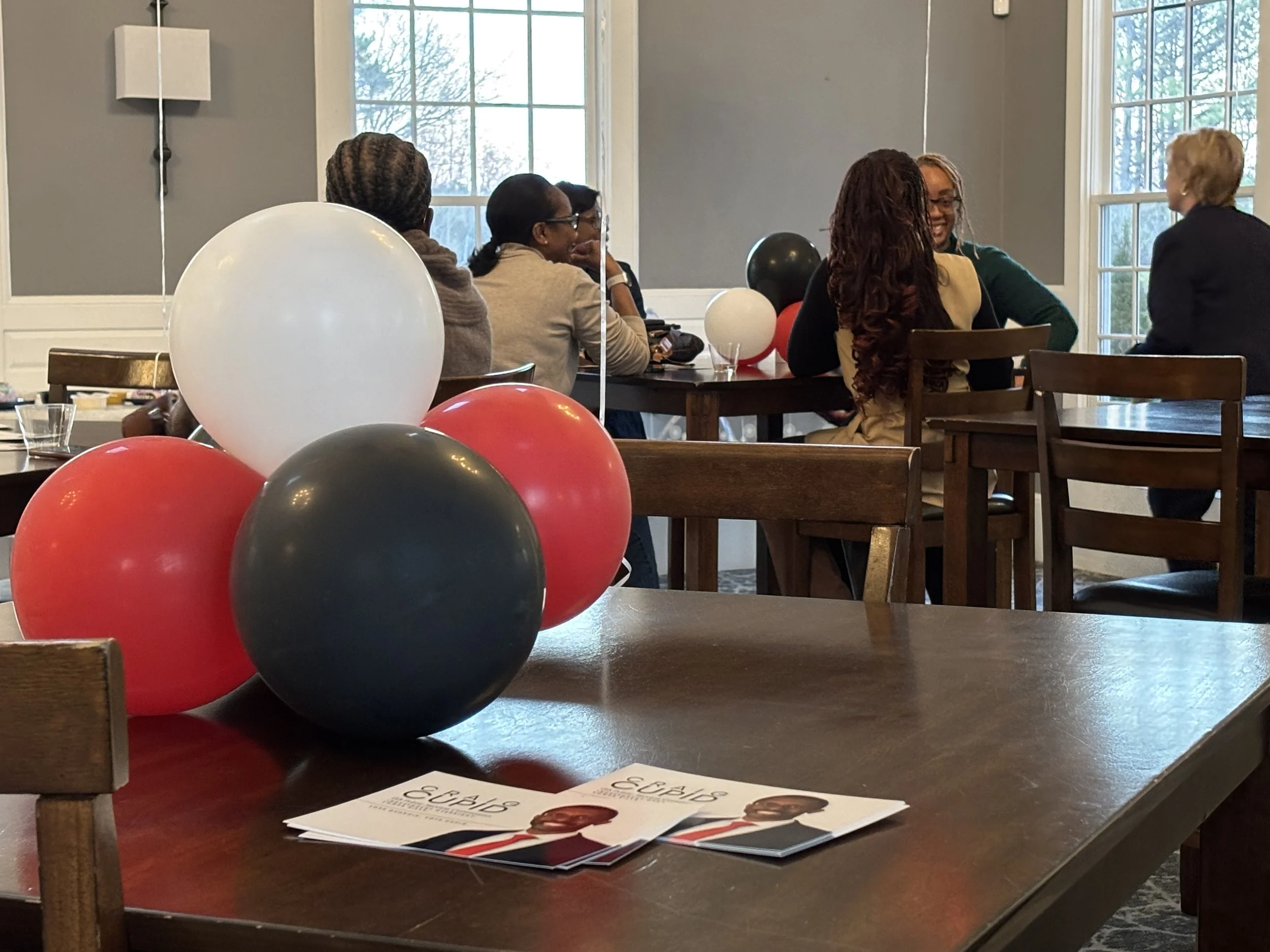 red black and white balloons on a dark wood table in the foreground with a group of people chatting in the background