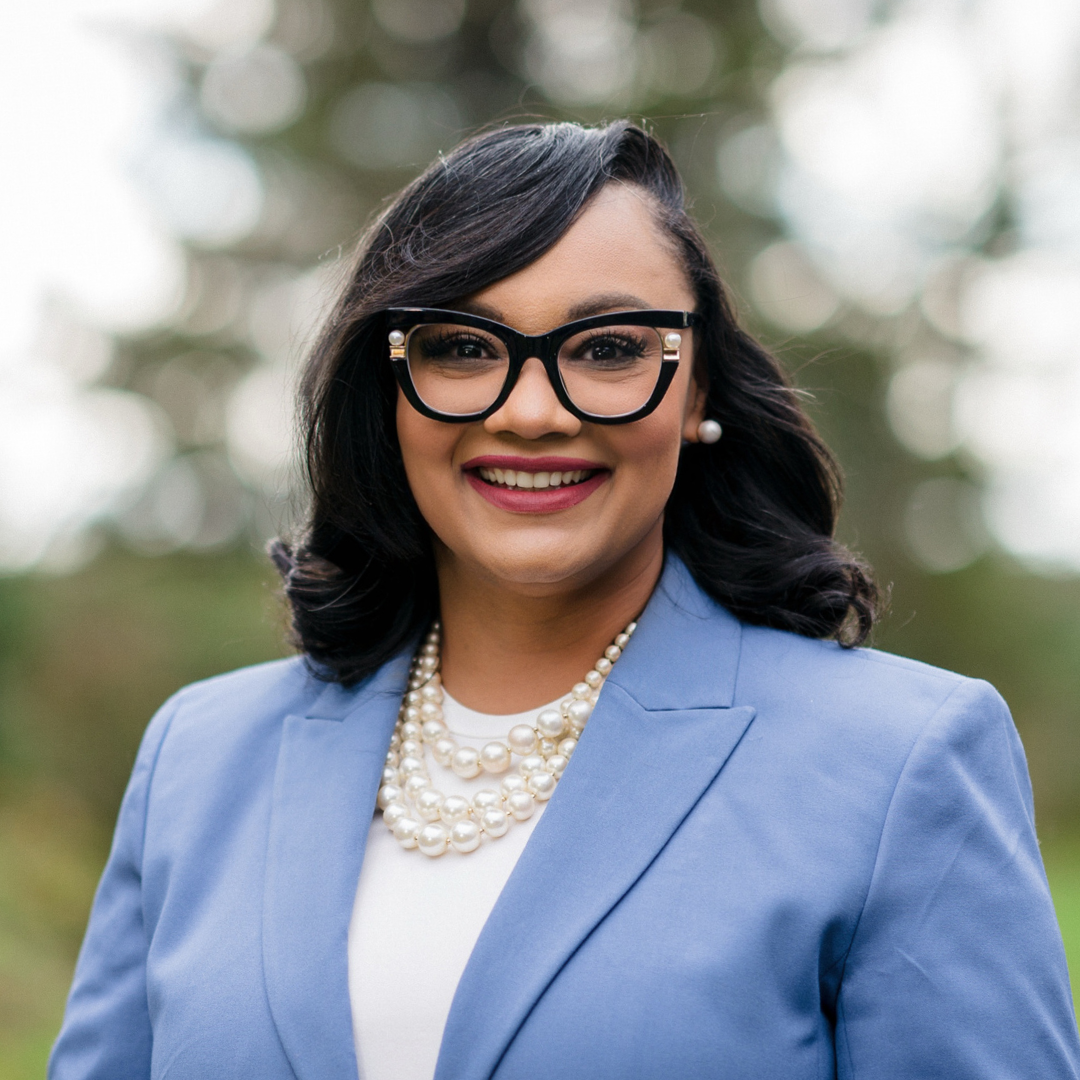 A woman with dark hair wearing glasses, a blue blazer, a pearl necklace, and pearl earrings, smiling outdoors.