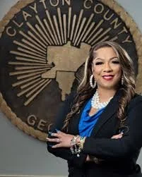 A woman with long dark hair smiling, wearing a black blazer over a blue top, standing in front of a circular emblem that says 'Clayton County Georgia' with a map of Texas and radiating lines.