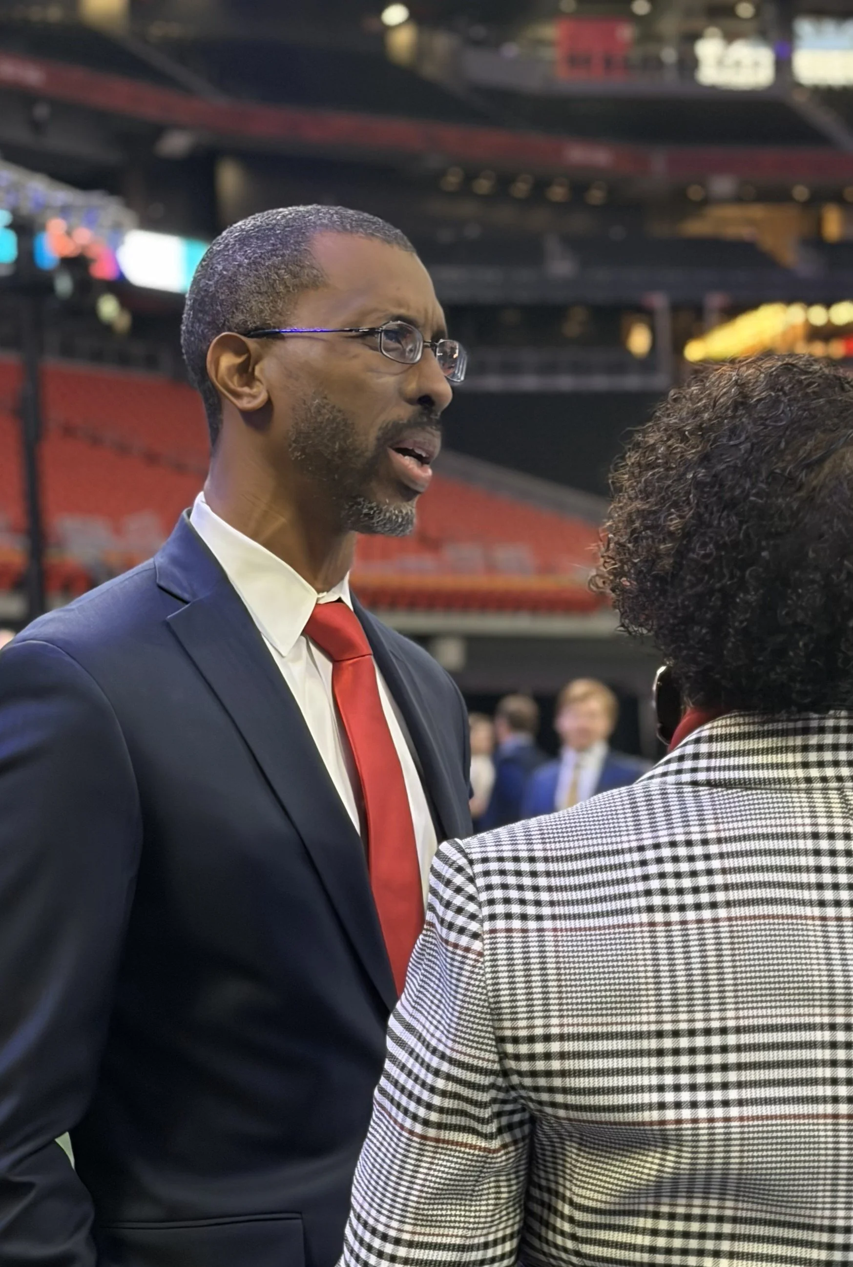 A man in a suit and red tie is talking to a woman with curly hair and glasses in a stadium or arena setting.