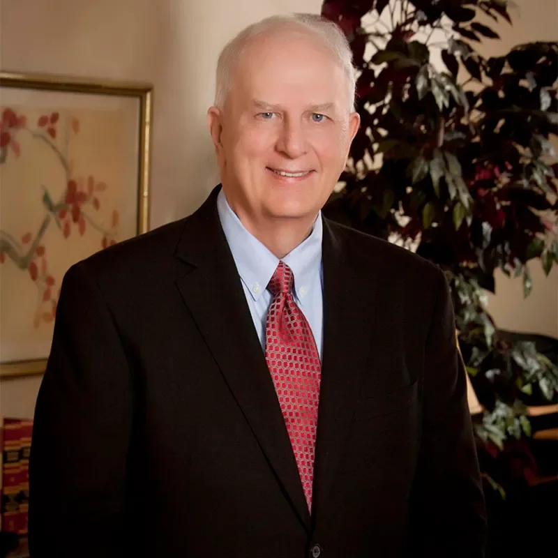 A middle-aged man with light gray hair wearing a black suit, light blue shirt, and red patterned tie standing indoors in front of a plant and framed artwork.