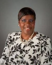 A woman with short black hair wearing a black on white floral shirt, smiling outdoors with a gray backdrop.