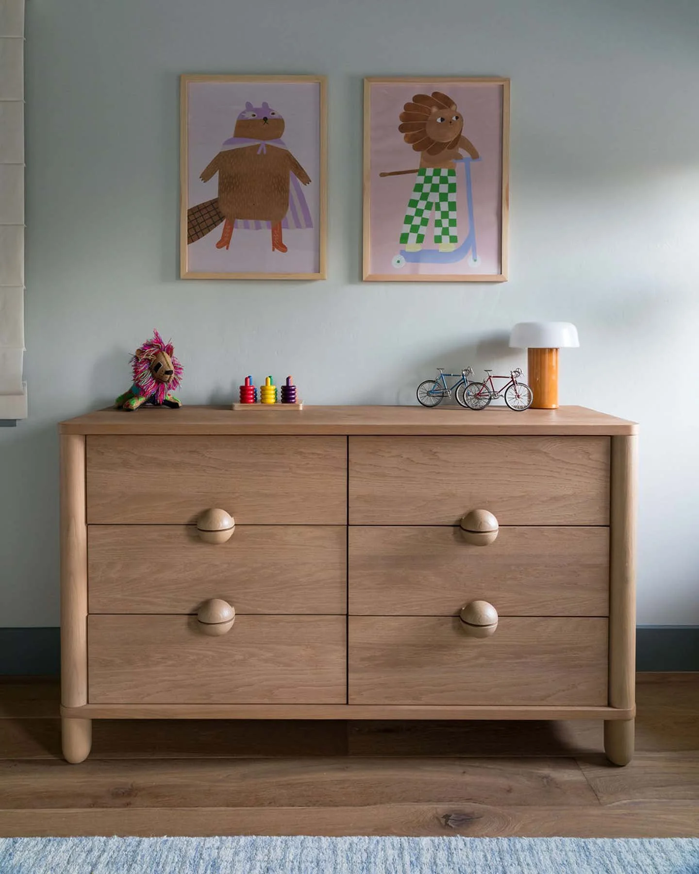 A wooden dresser with six drawers, simple round knobs, standing on a wooden floor. On top, there is a doll with pink mane, four small colorful stacking toys, two miniature bicycles, and a white and wooden table lamp. Two framed kid's artwork featuring characters are hung on the light green wall above the dresser.