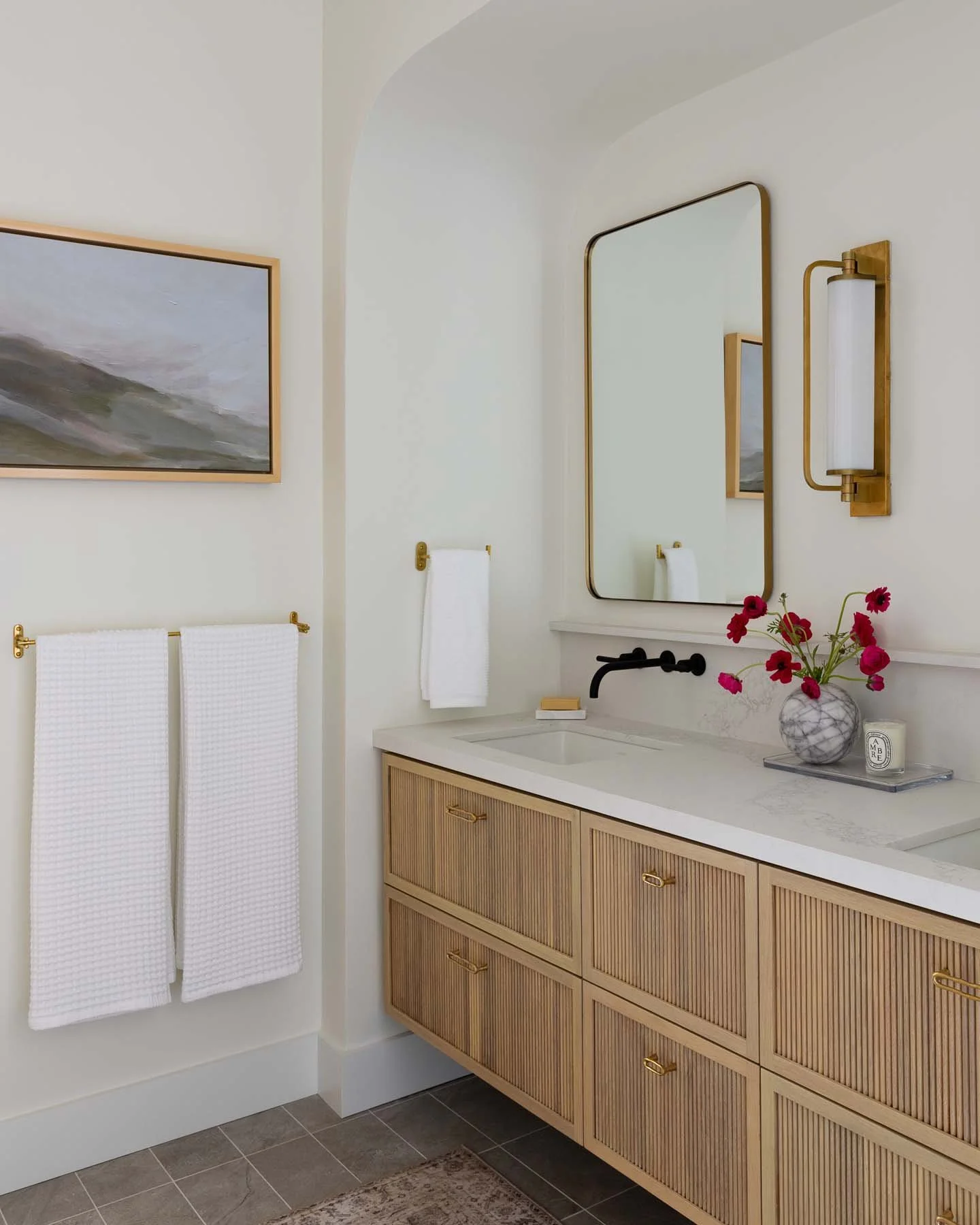 Bathroom with a wooden vanity, oval mirror, wall-mounted light fixture, white countertop, pink flowers in a vase, and white towels.