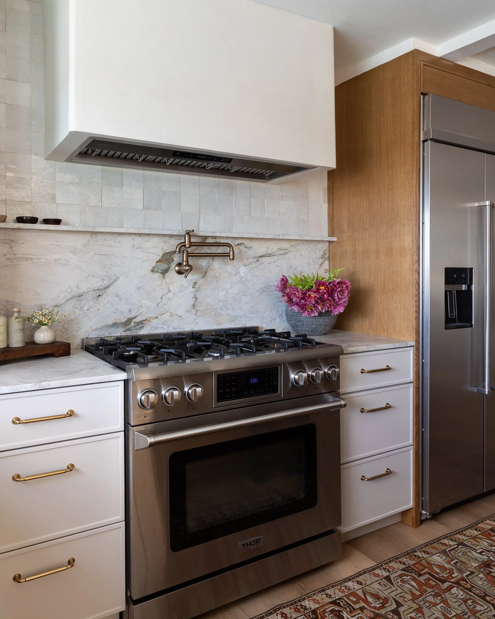 A modern kitchen with a stainless steel oven and stovetop, white cabinets with gold handles, a marble backsplash, a pot filler faucet, and a decorative potted flower on the countertop.