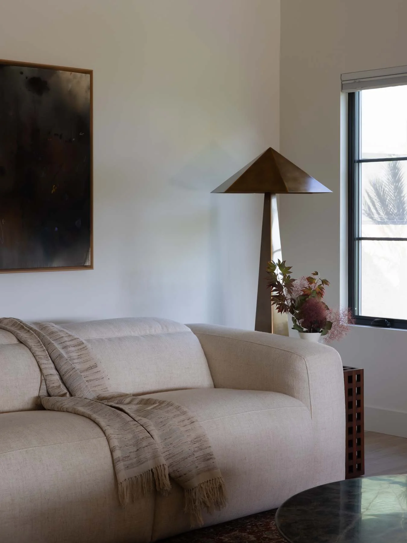 Living room with a cream sofa, a beige throw blanket, a wooden pyramid-shaped lamp, a window with a view of plants outside, and a flower arrangement on a side table.