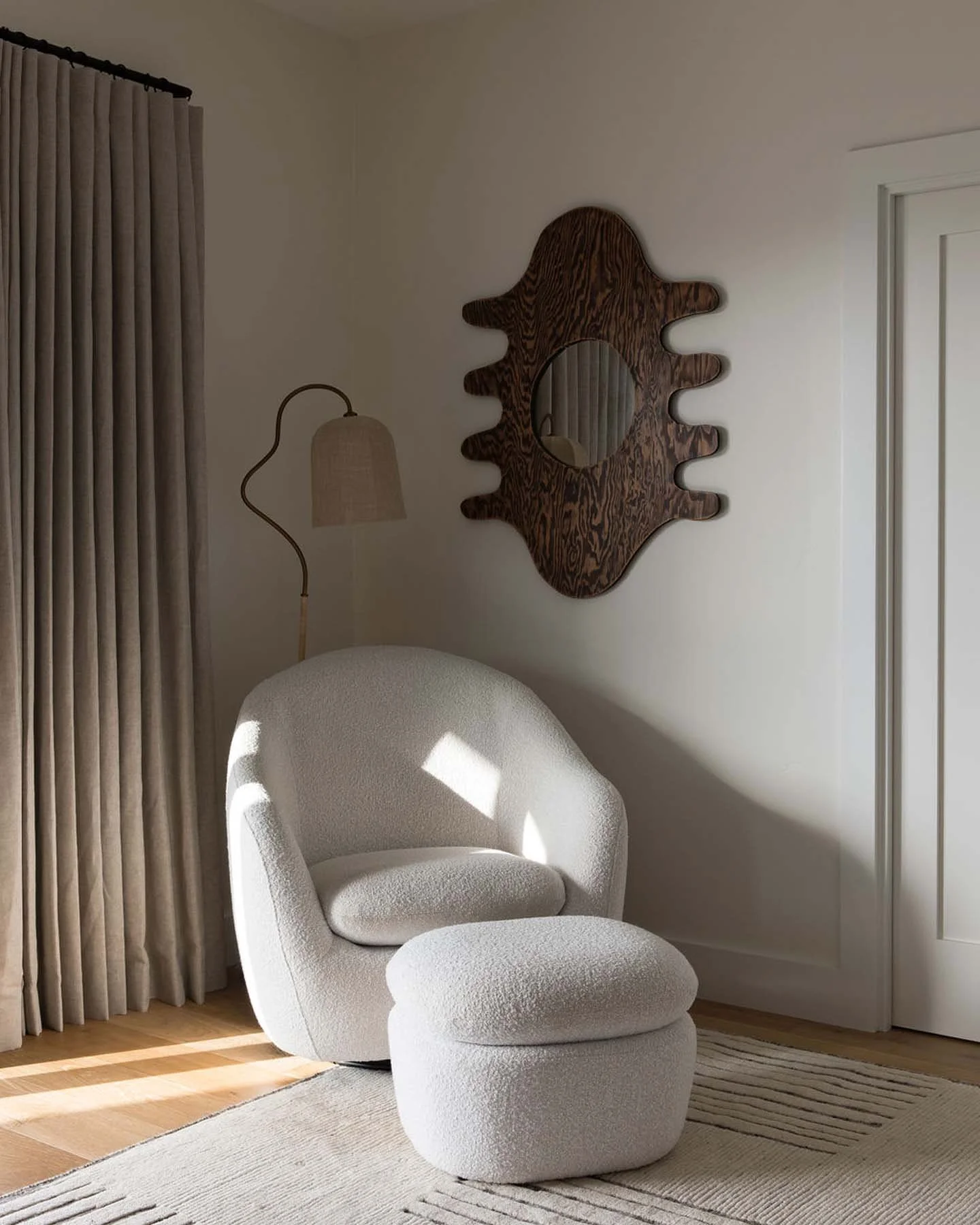 A white textured armchair with a matching ottoman, beside a wall with a decorative wooden mirror and a curved floor lamp, in a well-lit room with beige curtains and wooden floor.