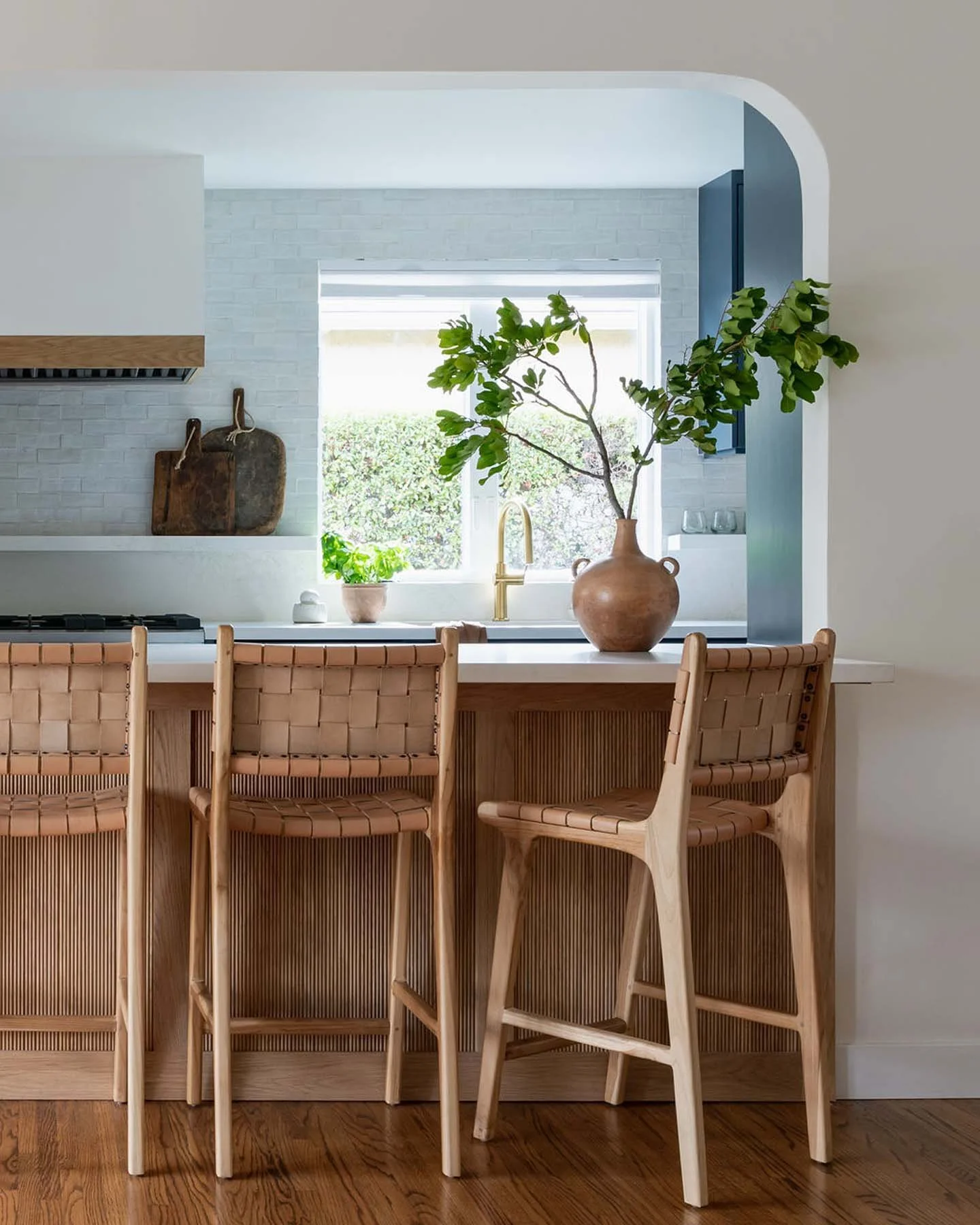 A modern kitchen with a white brick wall, a window with a view of greenery, wooden kitchen accents, a large vase with a leafy green plant on the counter, and a kitchen island with four woven bar stools.