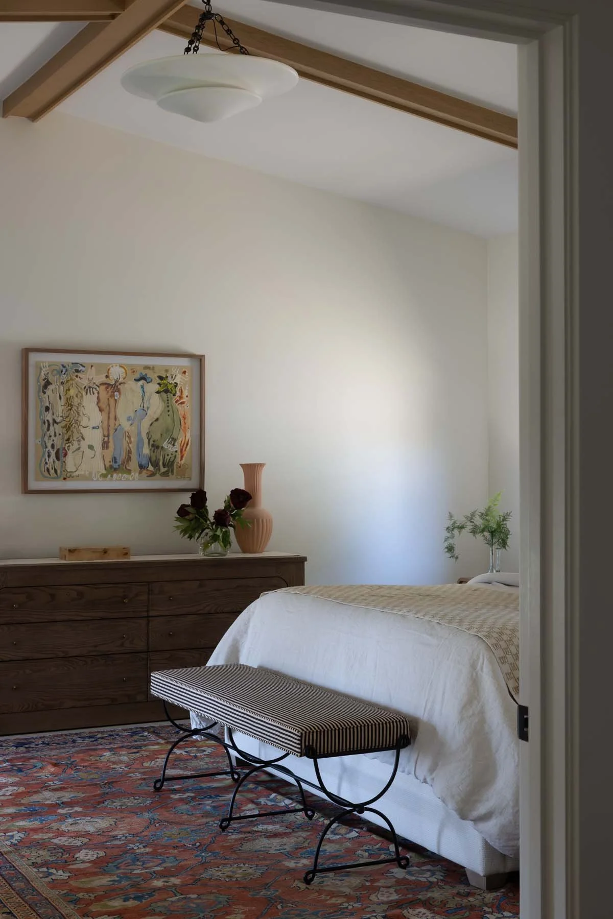 A bedroom with a white bed, a wooden dresser with a vase of dark red roses, a pink vase, and a framed abstract art piece on the wall. There is a small bench with a striped cushion in front of the bed, and a patterned area rug on the floor.