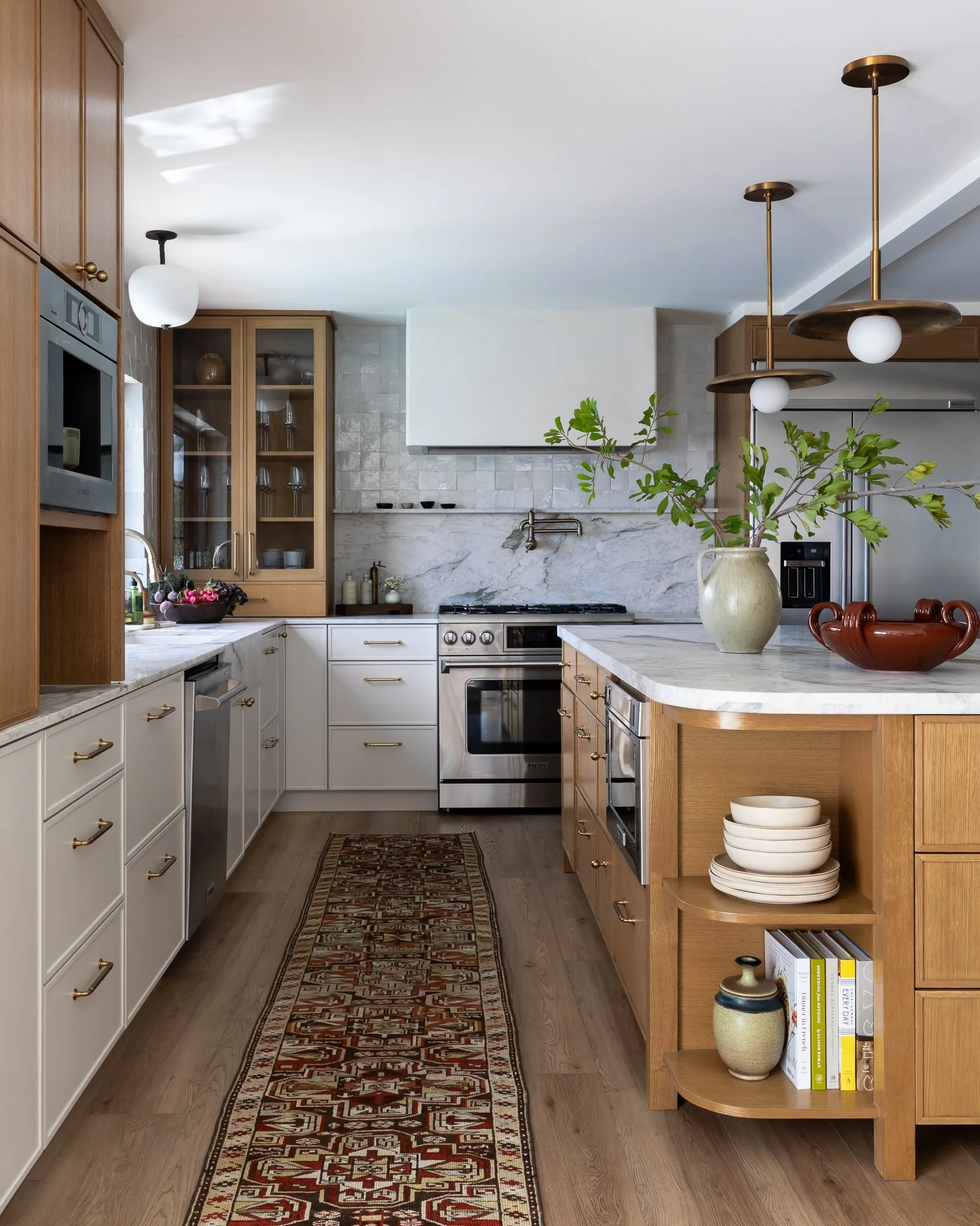 Modern kitchen with white and wooden cabinets, stainless steel appliances, marble countertops, a patterned runner rug, and decorative vases and books on open shelves.