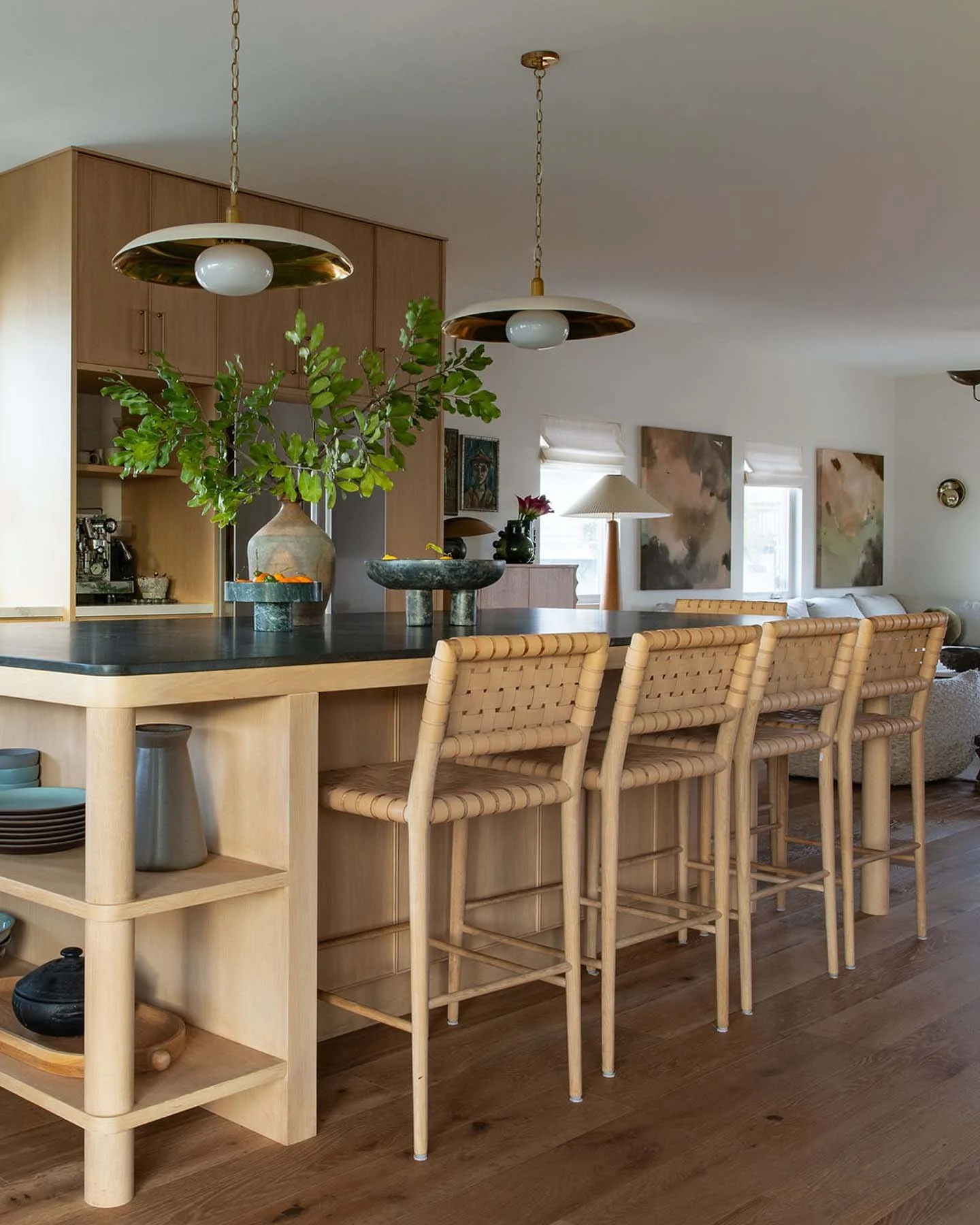 Open-concept living space with a kitchen island featuring a black countertop, light wood cabinetry, and modern pendant lights hanging from the ceiling. There are wicker bar stools along the island, and decorative vases with green foliage and flowers on the countertop. The background includes a sitting area with abstract wall art, a white sofa, a side table with a lamp, and a window with white blinds.
