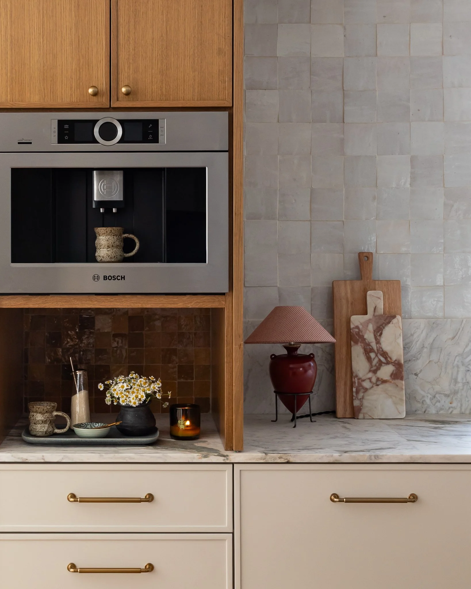 Interior of a modern kitchen with wooden cabinets, a Bosch coffee machine, decorative cups, a small vase with flowers, candles, and marble cutting boards on a countertop. The background features a tiled wall with neutral colors.