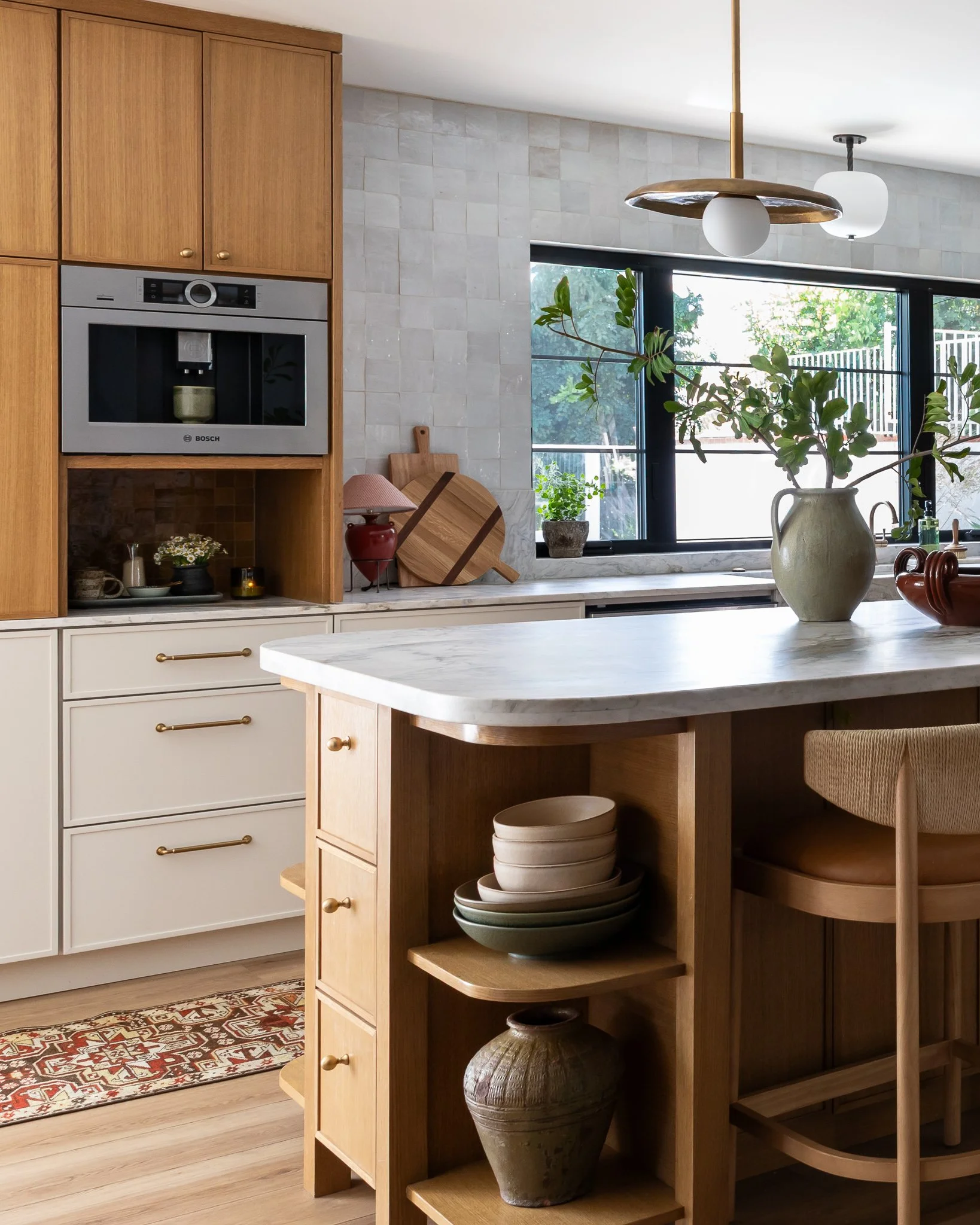 Updated kitchen featuring new cabinets, a marble countertop, and a large window with plants outside. There is a wooden open shelf with pottery and bowls, a leafy plant in a white vase, and a patterned rug. Designed by Vivo Design.