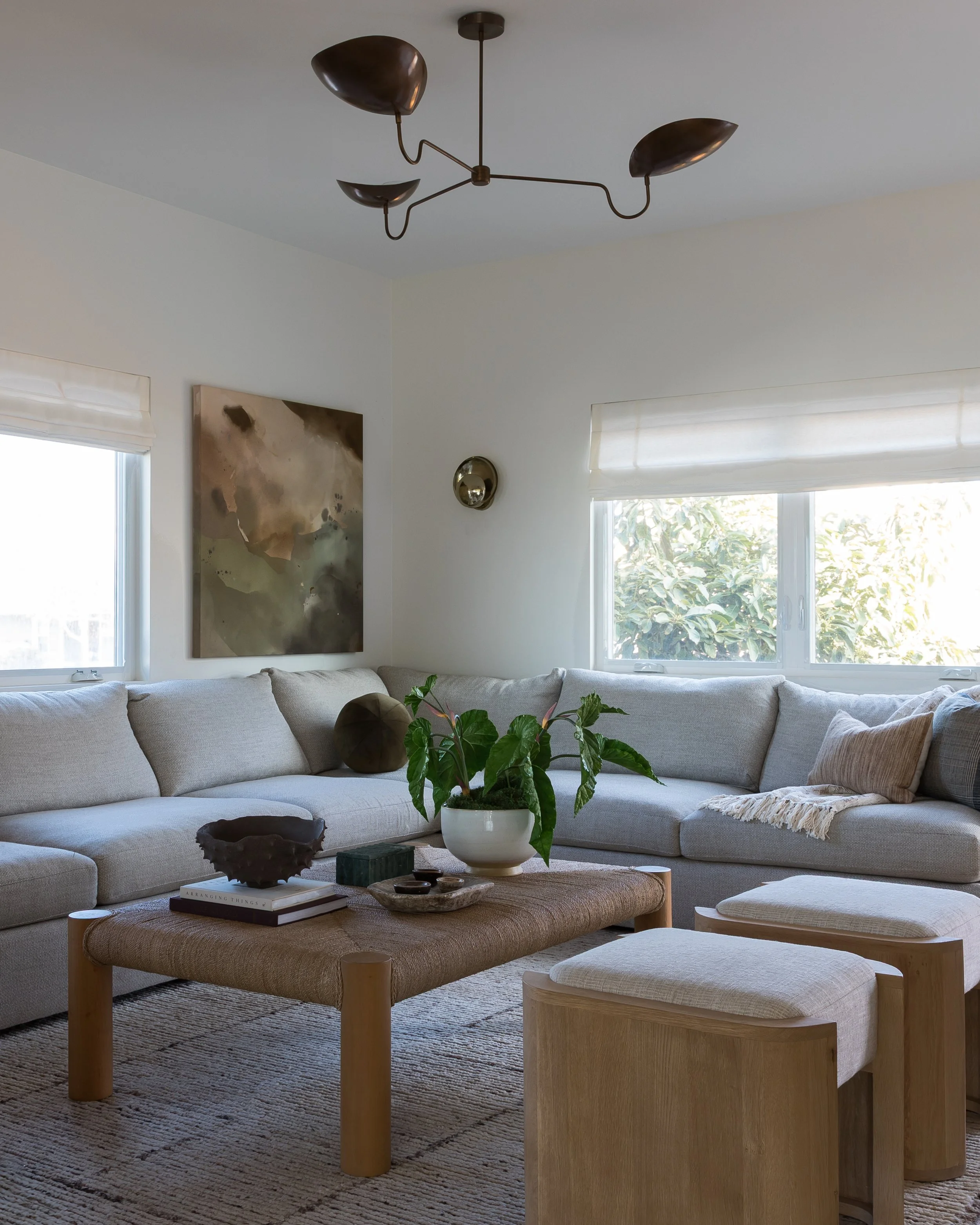 A cozy living room with a built-in white sectional sofa, a beige ottoman with a round top and wooden legs, a wooden stool with a matching cushion, and a large window with white blinds allowing natural light. The room features a modern chandelier, a wall painting, and a indoor plant.