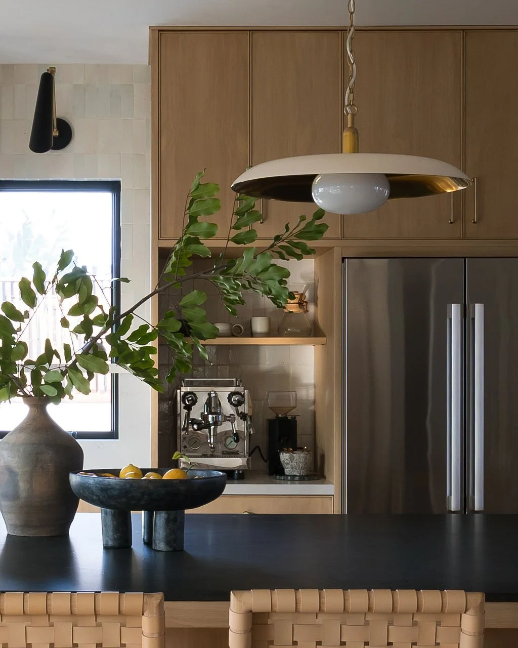 A modern kitchen with wooden cabinets, a large stainless steel refrigerator, a black countertop, a pendant light, a vase with green leaves, a bowl with lemons, and an espresso machine.