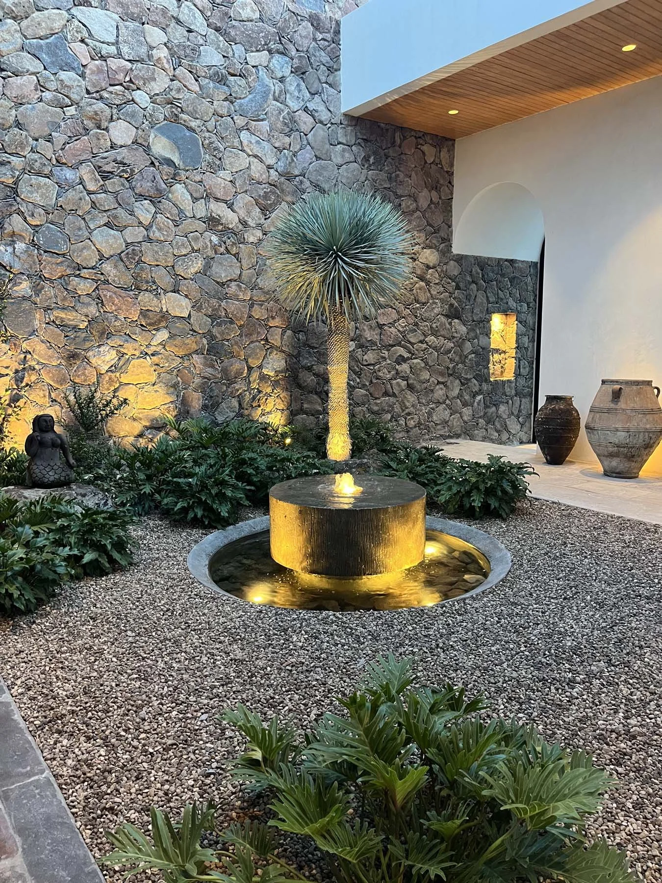 A modern outdoor courtyard with a stone wall, a palm tree, and decorative pots. There is a fountain with yellow lighting, plants, and a small Buddha statue, creating a tranquil ambiance.