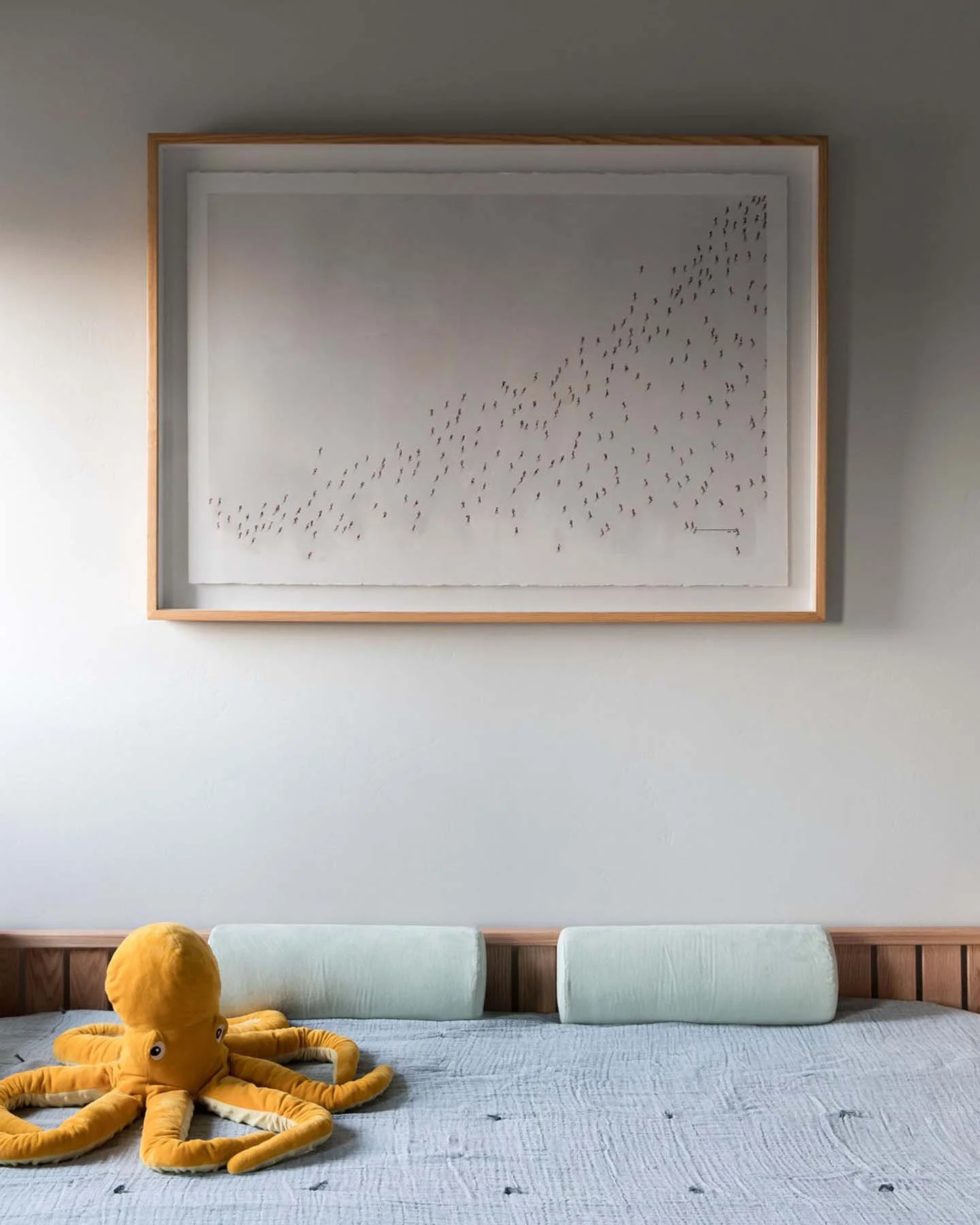 A bedroom with a framed art piece hanging on a light gray wall, a bedsheet with small patterns, a plush yellow octopus toy on the bed, and three green cushions on the headboard.