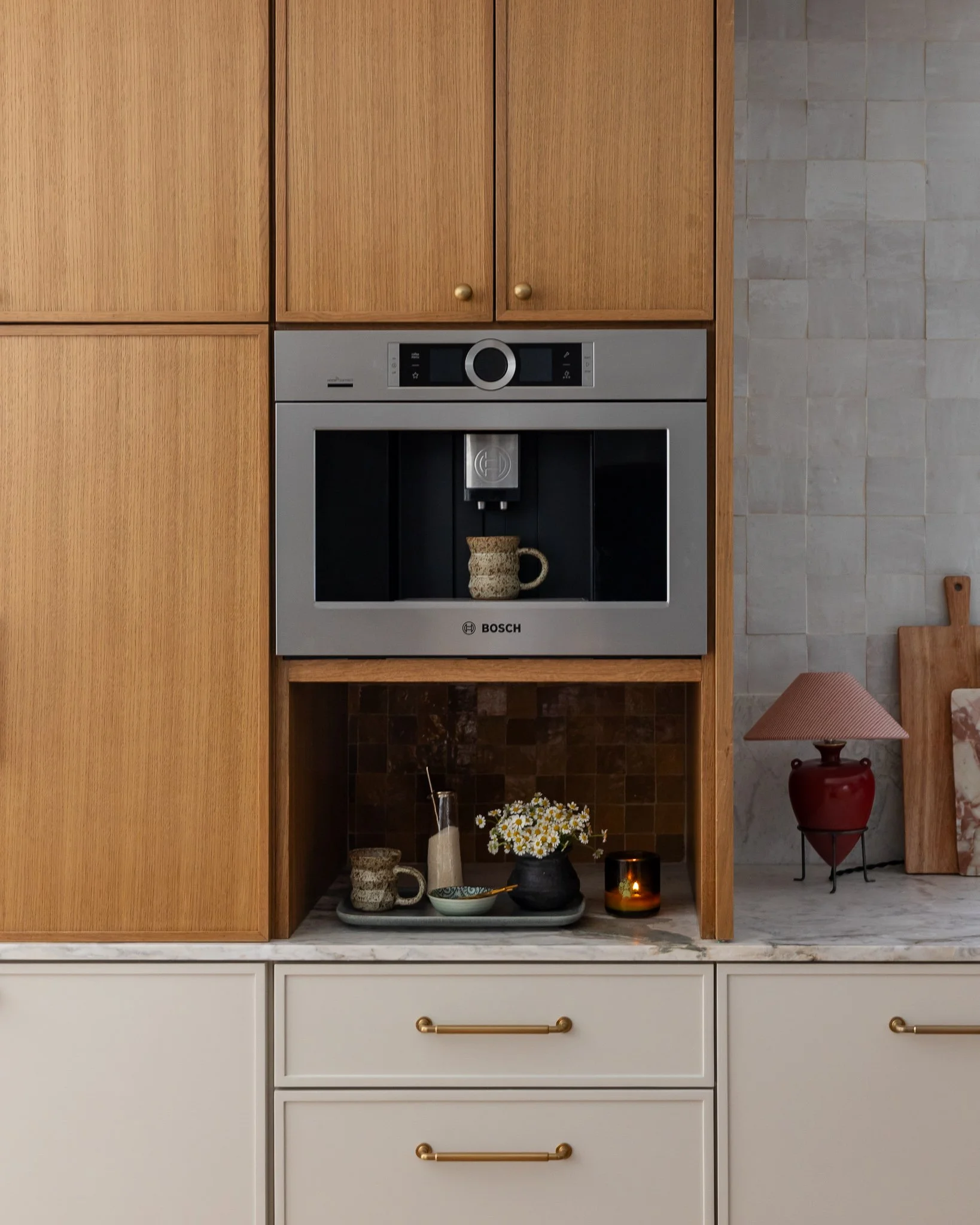Kitchen cabinets with built-in oven, open shelf with decorative dishes, flowers, candles, and a red vase on marble countertop.