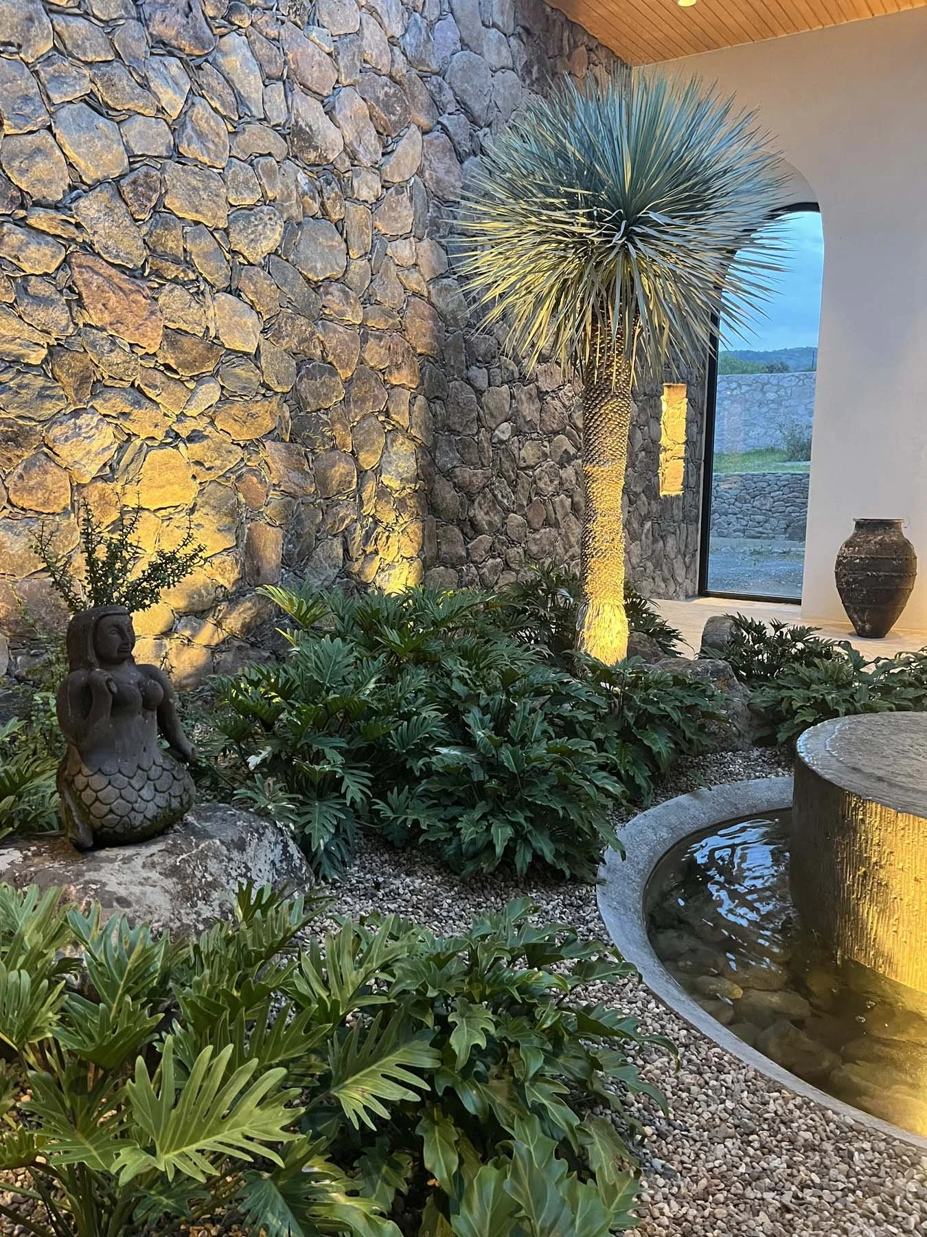 Indoor garden with stone wall, a tall desert plant illuminated by warm lighting, surrounded by lush green plants, a small mermaid statue, and a water fountain with built-in lighting.