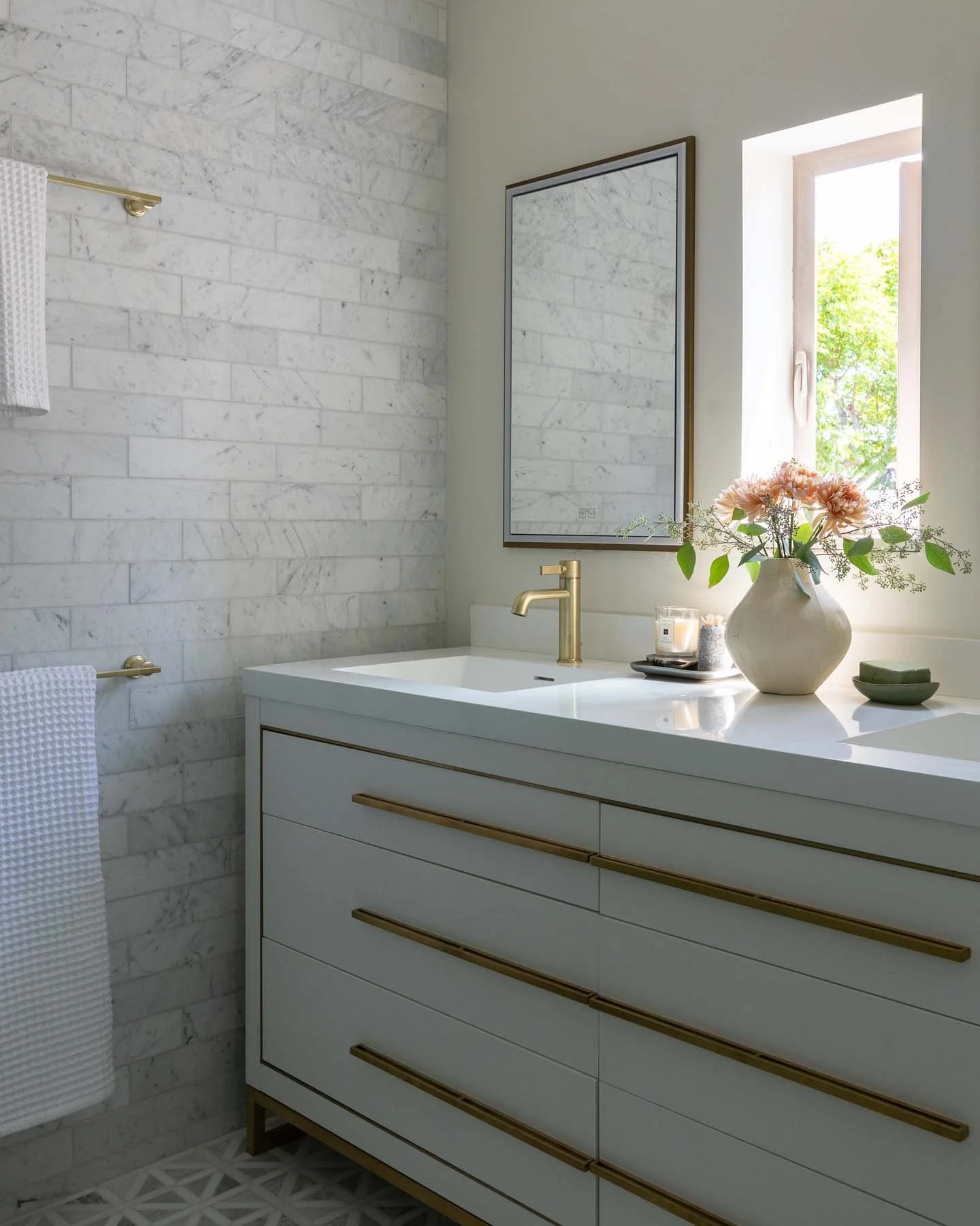 A bathroom vanity with six drawers, gold handles, and a white countertop. On the countertop, there is a large white vase with pink flowers and green leaves. Next to the vase, there is a candle in a glass container and a small dish. A mirror is mounted above the vanity, reflecting the marble wall and window. Natural light is coming through a window with a view of green trees outside.