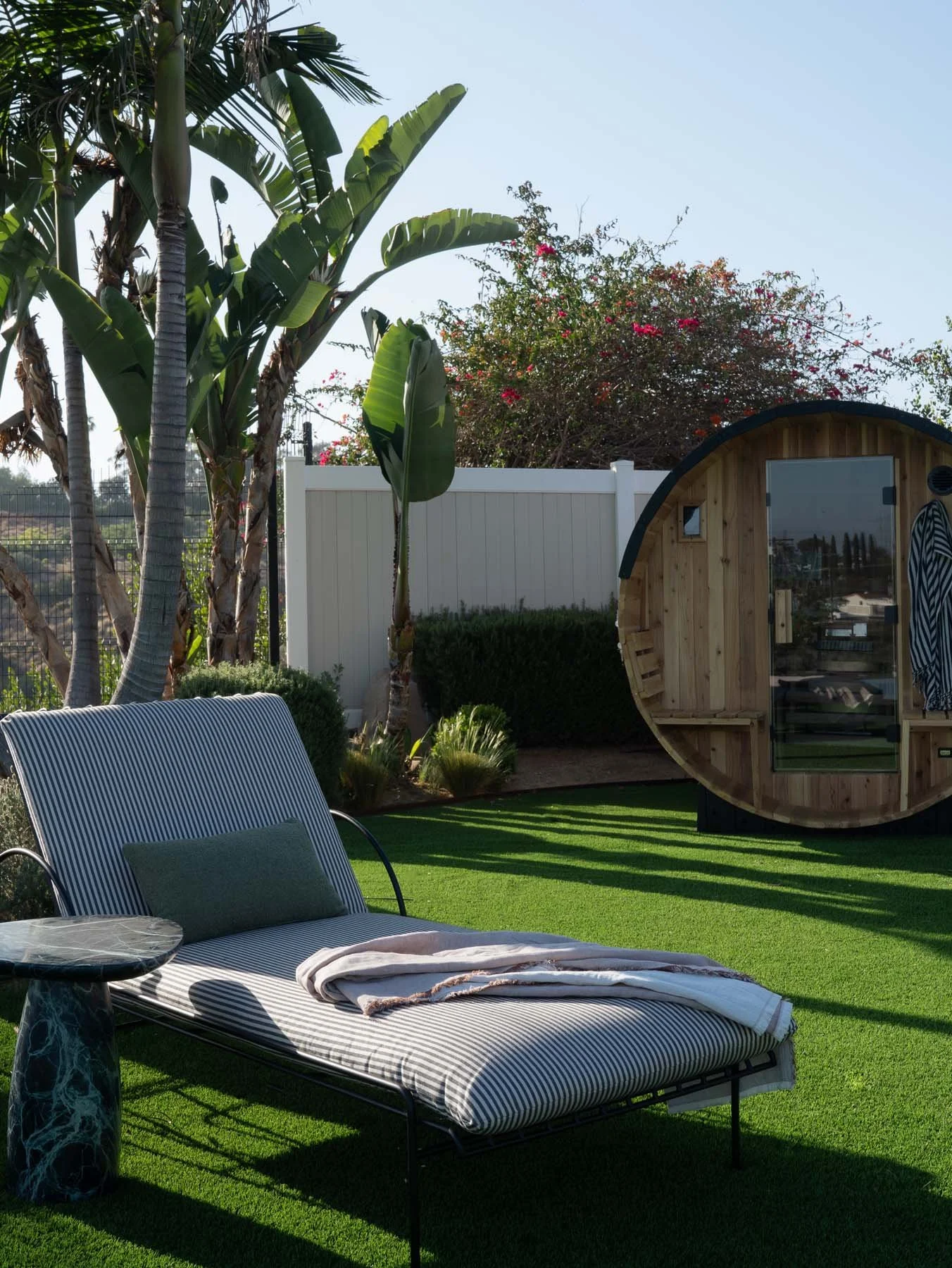 An outdoor backyard scene with a striped lounge chair with a pillow and a blanket, a small round table with a marble top, palm trees, a white fence, a shed with a curved roof made of wood and glass, and a lawn with artificial grass.