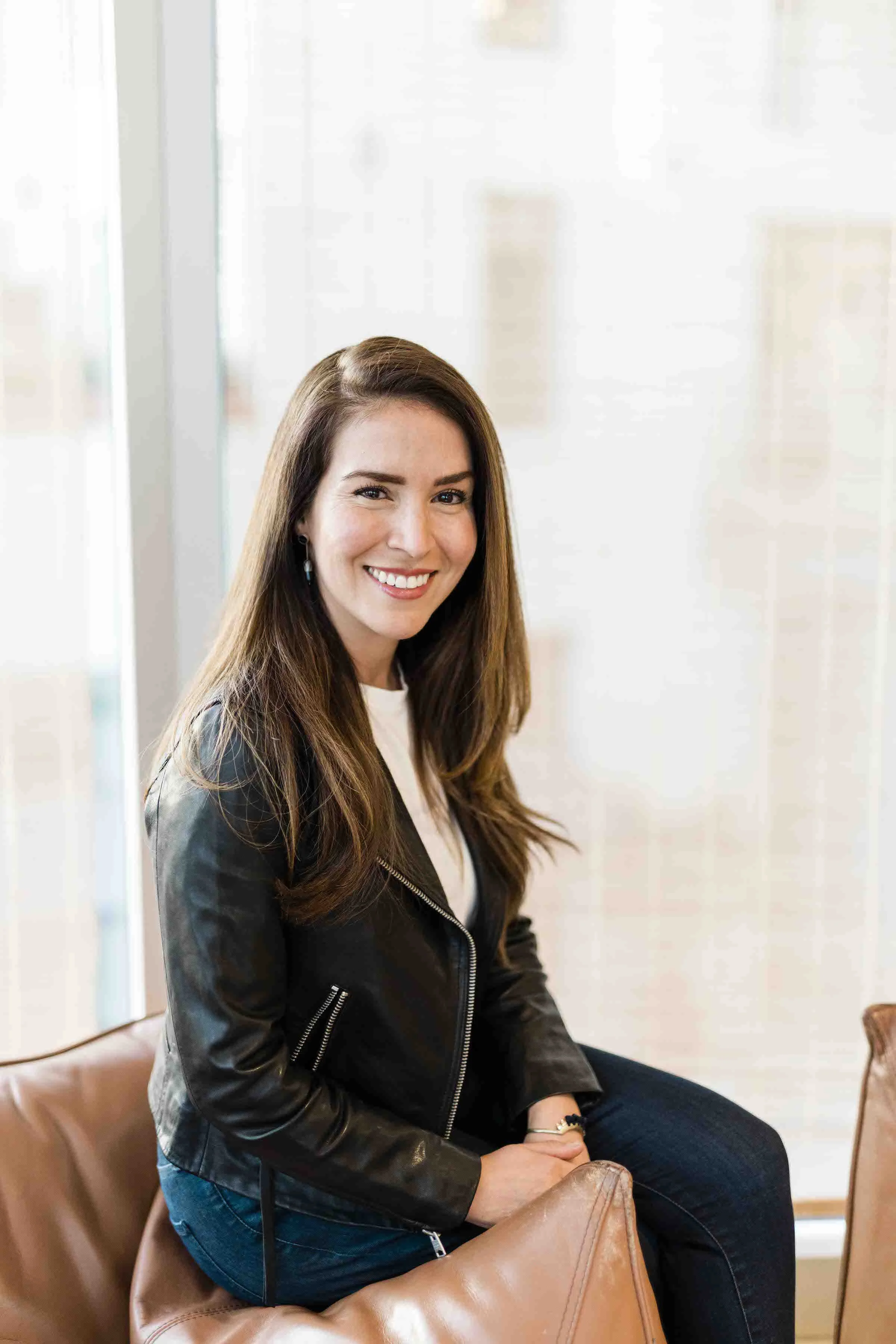 A woman with long brown hair smiling, sitting on a brown leather chair in a bright room with white walls and windows, wearing a black leather jacket and dark pants.