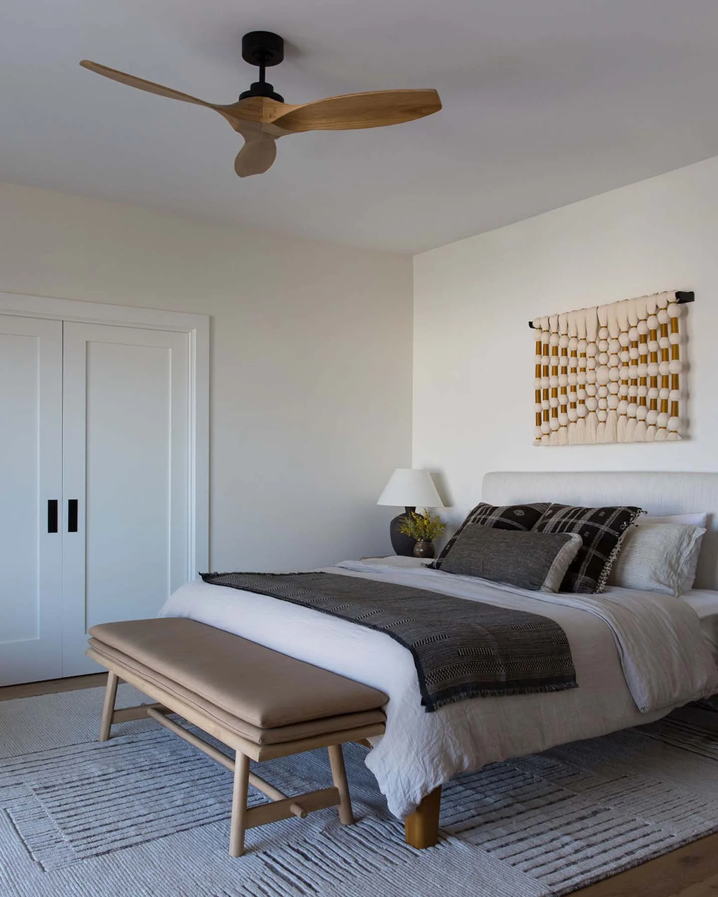 A bedroom with a white bed, beige bench, black and white bedding, and pillows. A wooden fan on the ceiling, artwork on the wall, window with white door, and a lamp on a nightstand.