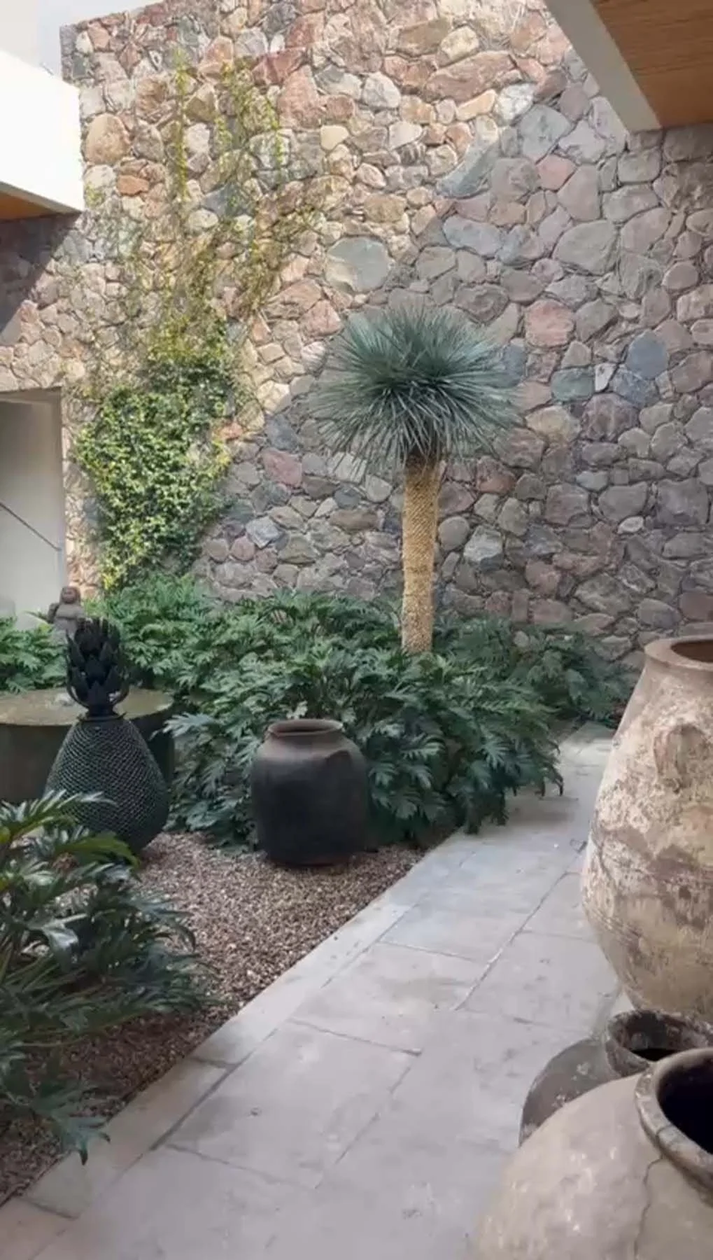 A courtyard with a stone wall in the background, a palm tree, green plants, and decorative pottery and vases.