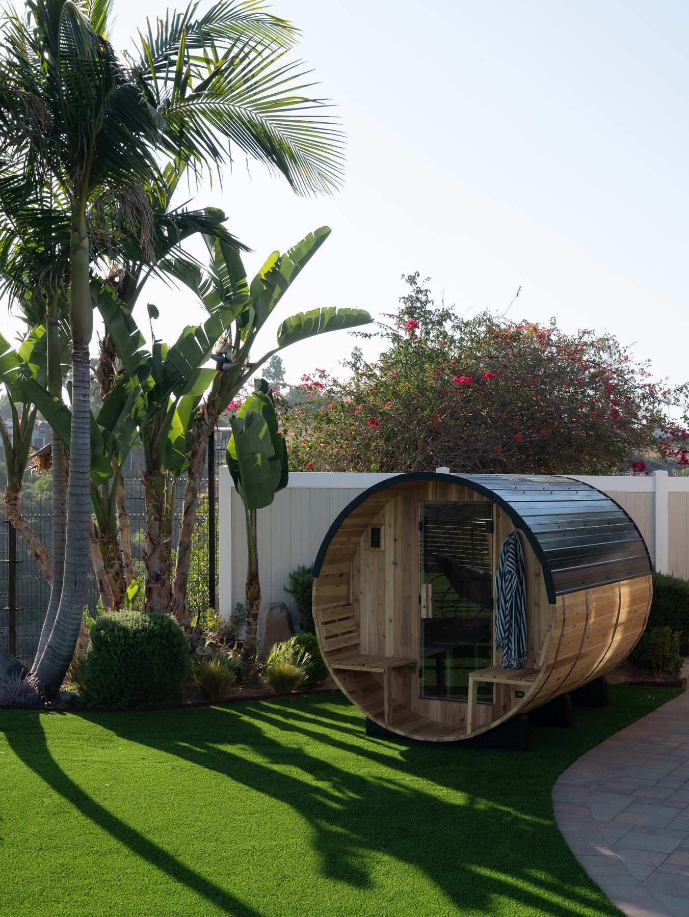 A backyard scene with a tall palm tree, blooming bushes, and a framed backyard with a small wooden round-shaped pod seating area on a green artificial grass lawn.