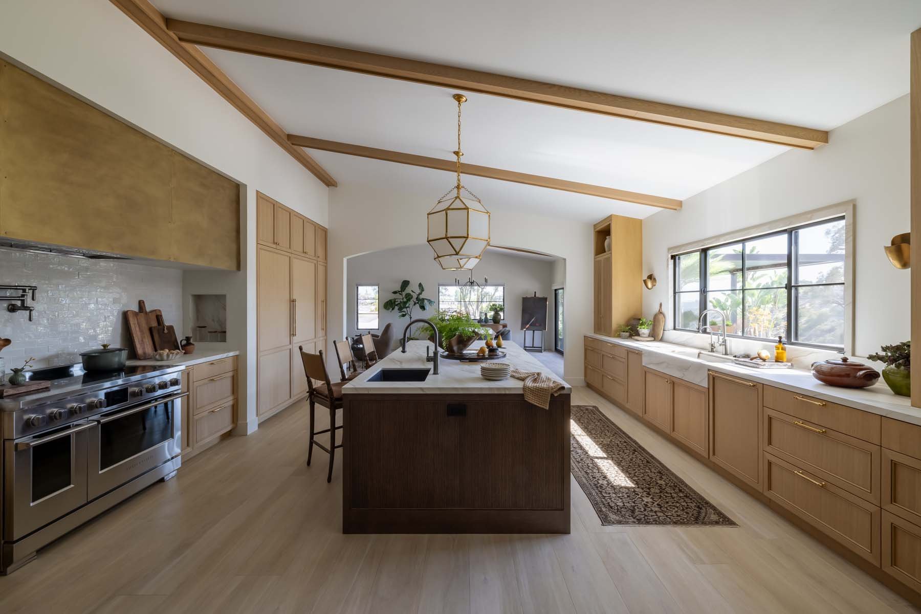 Bright, spacious kitchen with light wood cabinets, stainless steel stove, large island with sink, and a row of windows letting in natural light. Decor features plants, bowls, and a patterned rug.