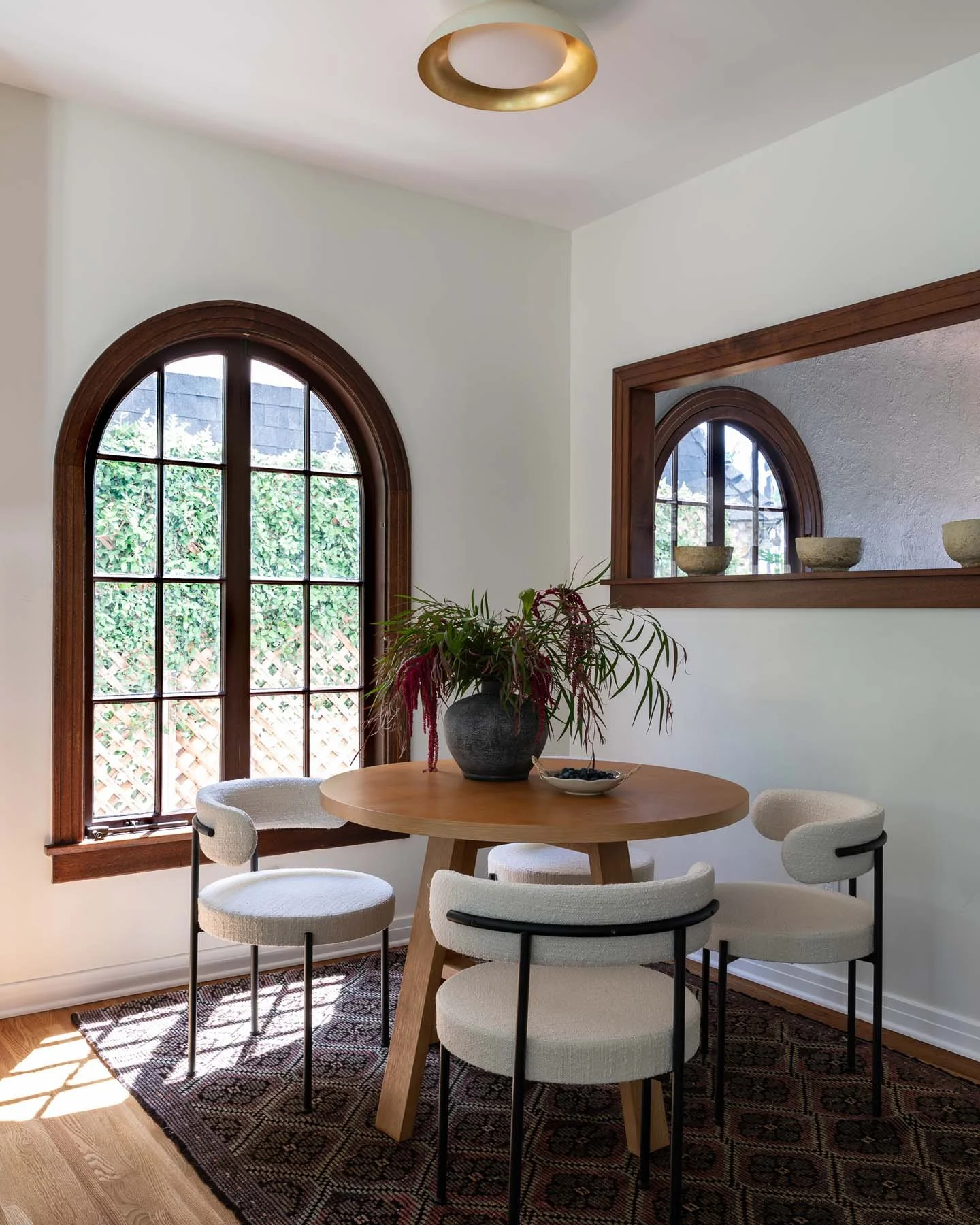 A dining room with a round wooden table, four white upholstered chairs, a potted plant centerpiece, a window with wooden trim, and a decorative mirror on the wall above the table. Sunlight streams through the window, casting shadows on a patterned rug.