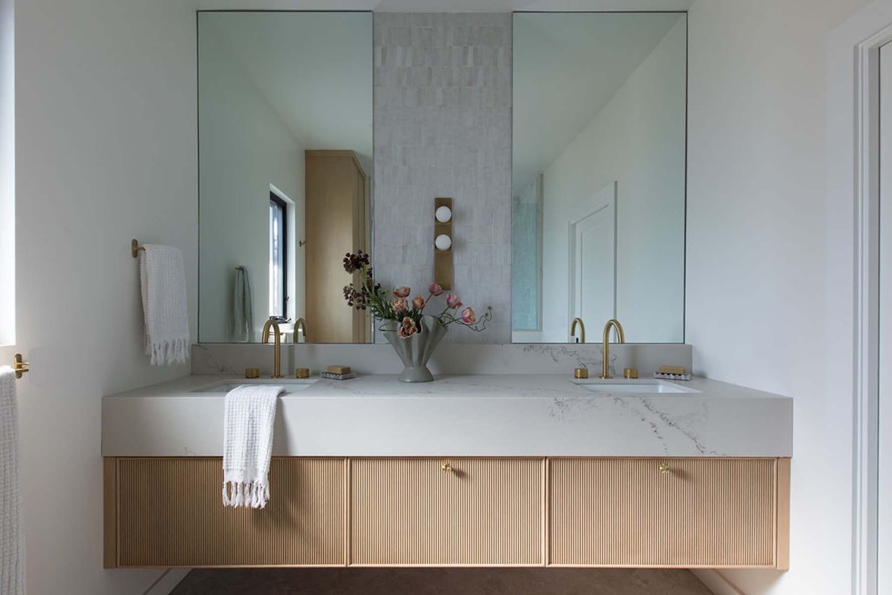 Modern bathroom vanity with two sinks, gold faucets, and a large mirror. A flower arrangement in a vase is placed between the sinks. White towels hang on gold towel bars, and the vanity has a wood finish with vertical grooves.