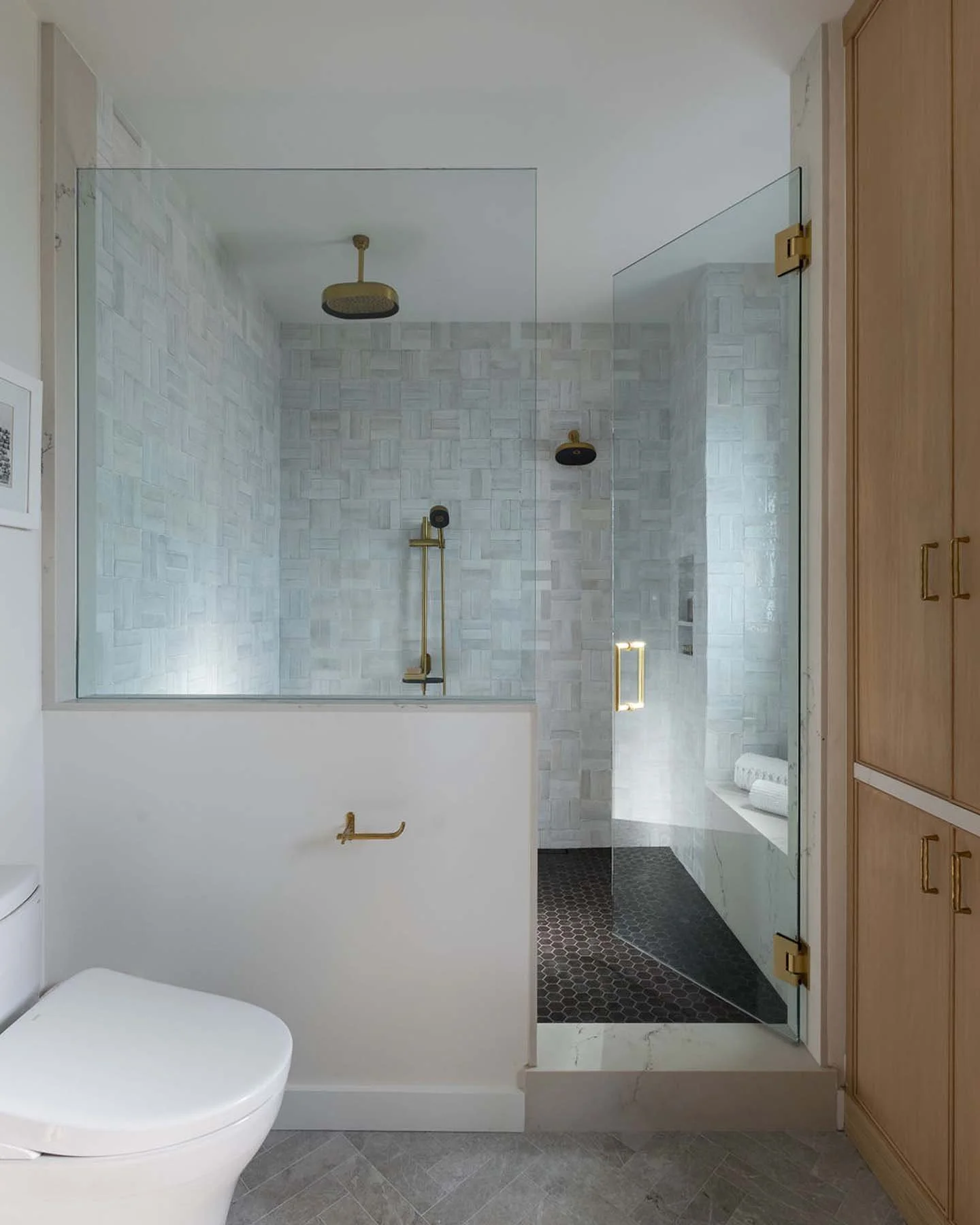 Modern bathroom with a walk-in shower featuring white textured tiles, black hexagonal floor tiles, brass fixtures, and a glass partition, with a white toilet and wooden cabinet.