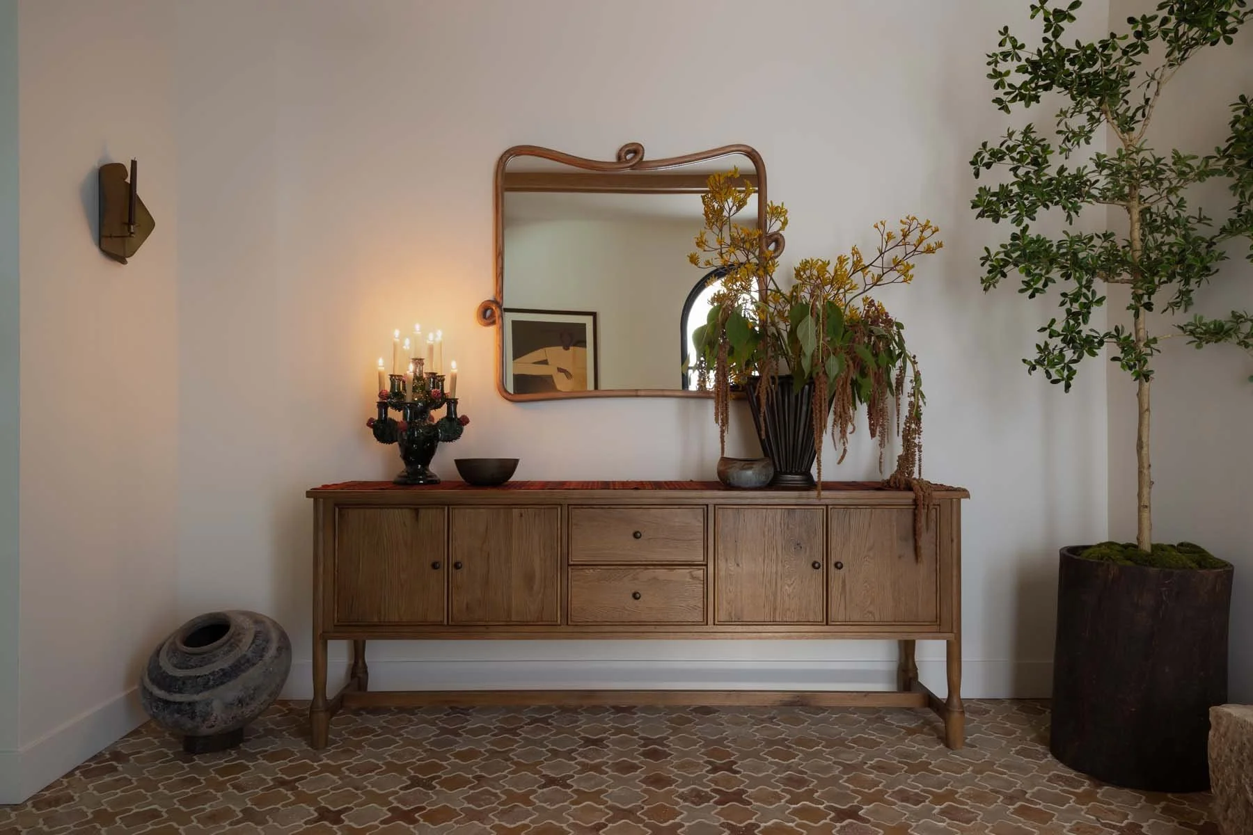 A wooden sideboard with three drawers and four cabinet doors, decorated with a black vase filled with yellow flowers, a black bowl, and a decorative candelabra with lit candles. A mirror with a wavy wooden frame hangs above the sideboard. To the right, there is a large potted plant with green foliage. On the left, on the floor, there is a round decorative object with a hole in the center. The room has beige patterned tile flooring and white walls.