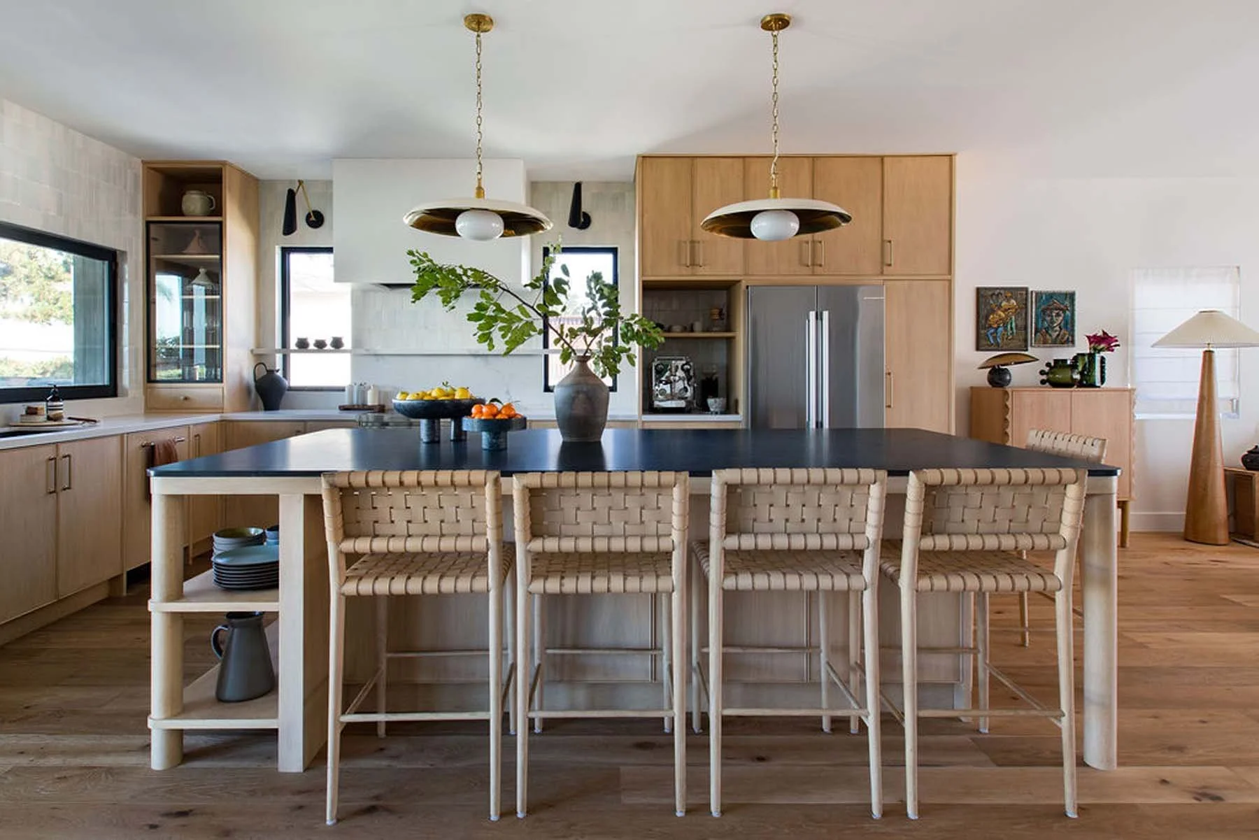 Modern kitchen with wooden cabinetry, black countertop island, and woven chairs. Decor includes a large vase with greenery, fruit bowls, artwork, and lamps.