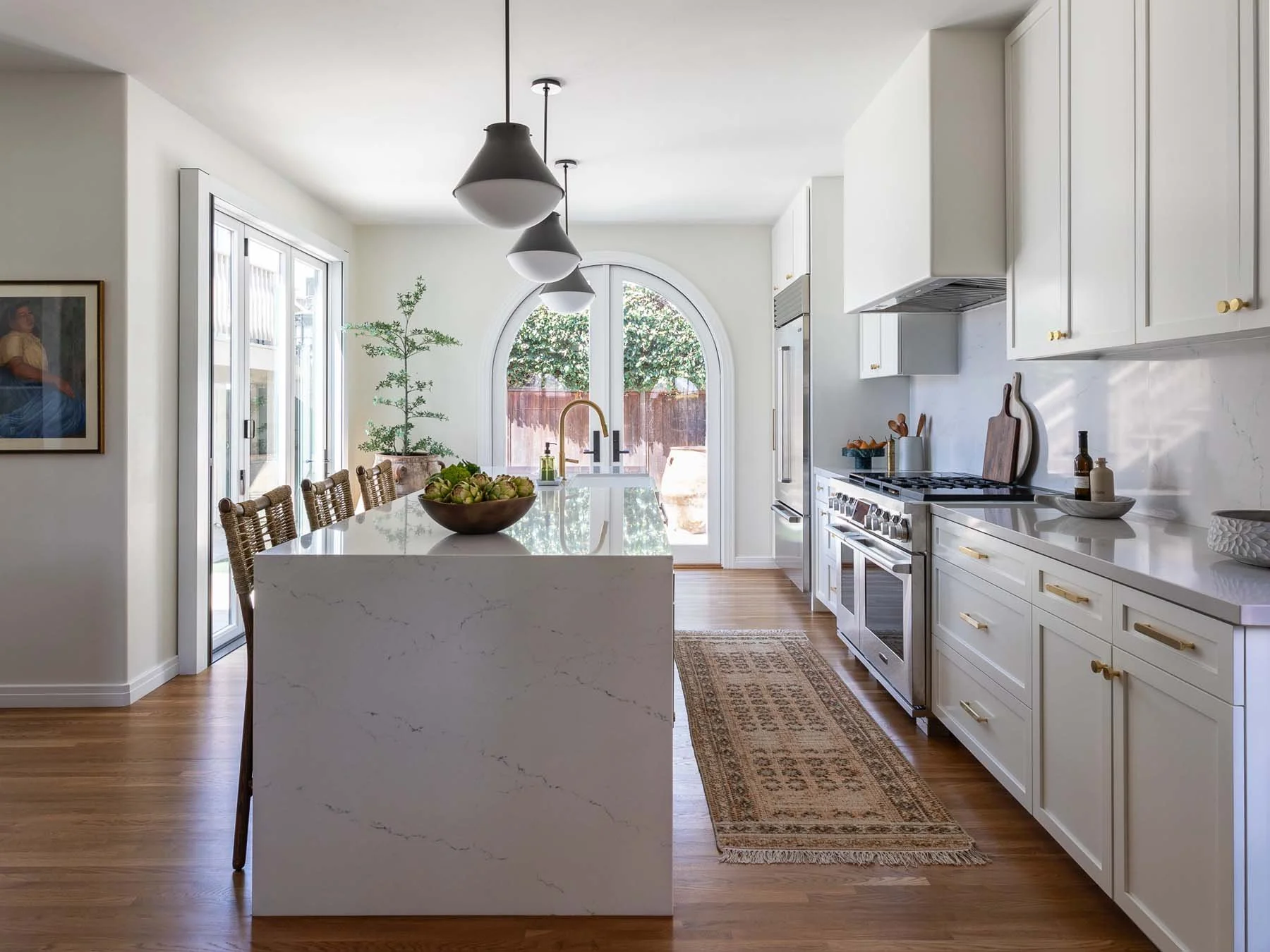 Modern white kitchen with wooden flooring, a marble island, and white cabinetry with brass hardware. Three black pendant lights hang above the island. A large window and sliding door let in natural light. Decor includes a bowl of artichokes on the island, wooden cutting boards, and a small potted tree near the window. There is a patterned rug on the floor.