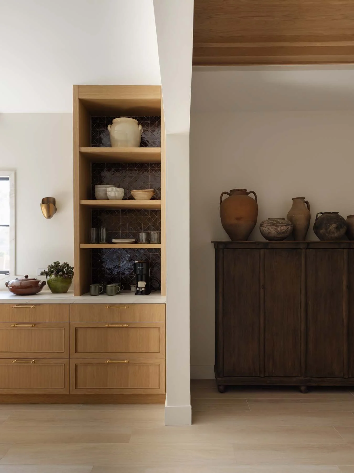 Interior view of a kitchen with a light wood cabinet, open shelves displaying dishes, a coffee maker, and cups, and a dark wooden cabinet with ceramic vases on top, against a white wall and light wood flooring.