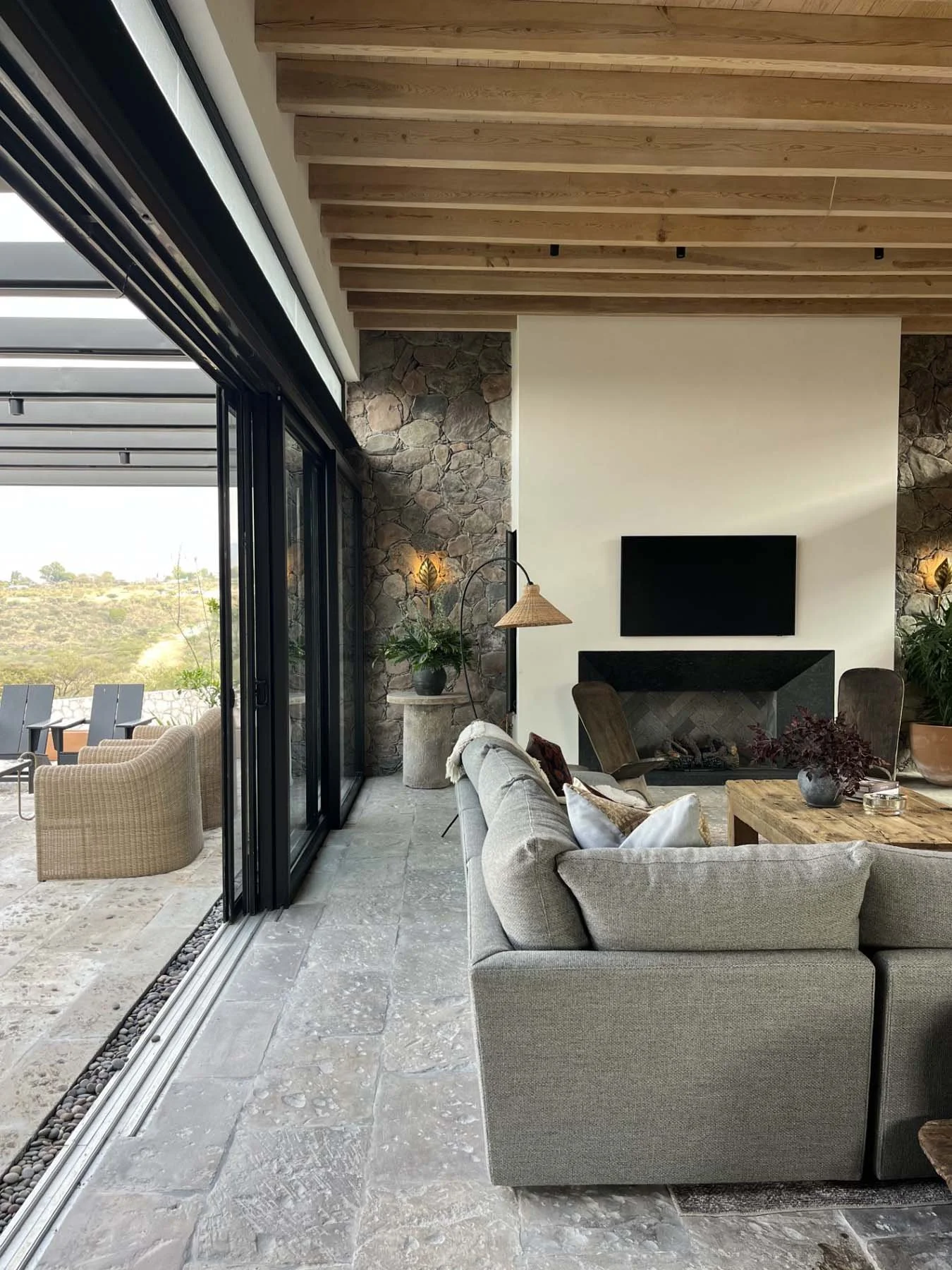 Living room with large glass sliding doors leading to an outdoor patio, stone fireplace, wooden ceiling beams, ceiling mounted lights, beige sofa, brown armchairs, coffee table with plants and decorative items, wall-mounted TV, and a view of a landscape outside.