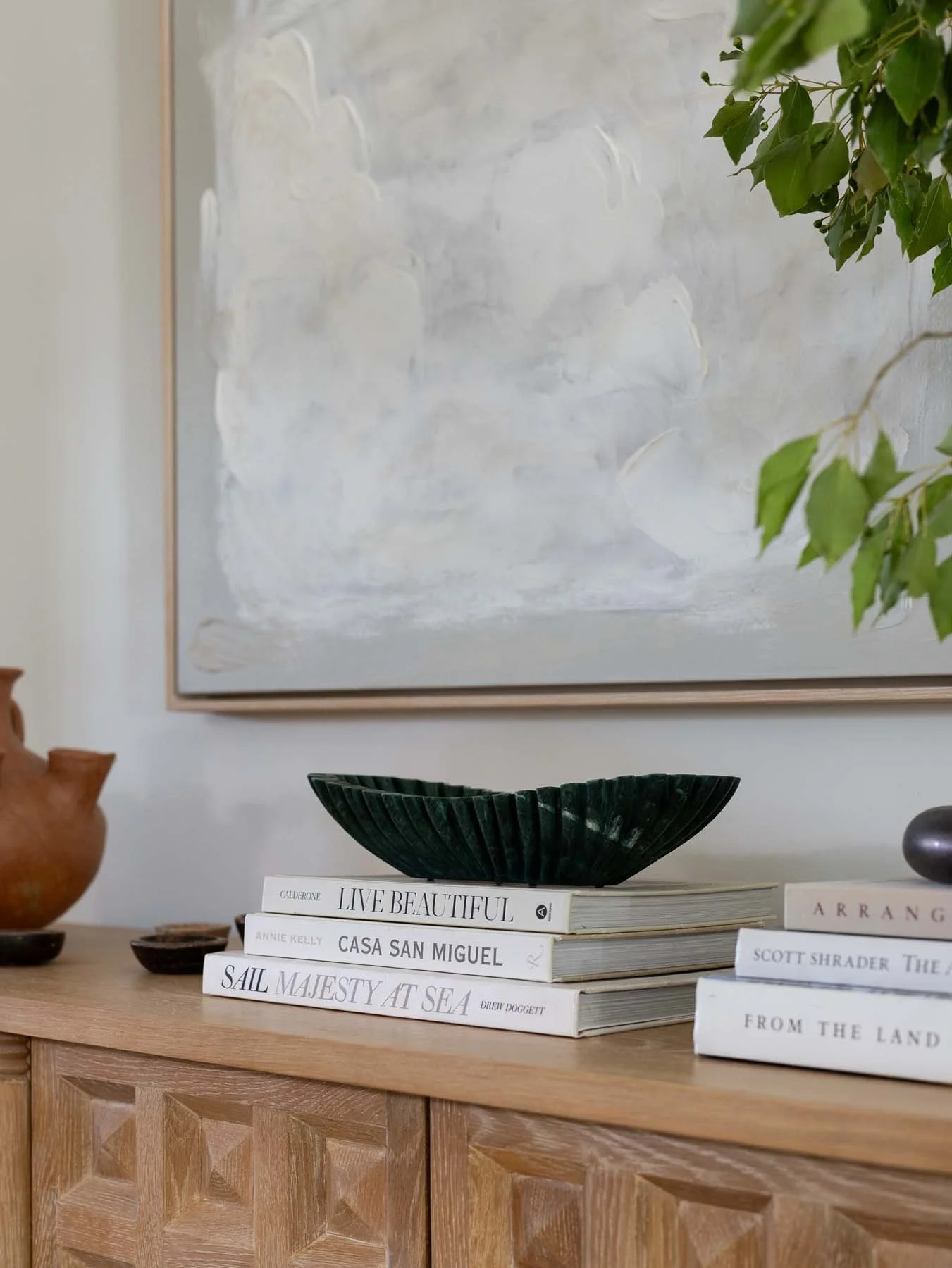 A wooden sideboard with a dark green decorative bowl on top, and a stack of books with titles such as 'Live Beautiful,' 'Casa San Miguel,' and 'Sail Majesty at Sea.' To the left, there is a pottery vase, and to the right, part of a black bowl and a gray object are visible. A large abstract painting with neutral tones hangs on the wall above.