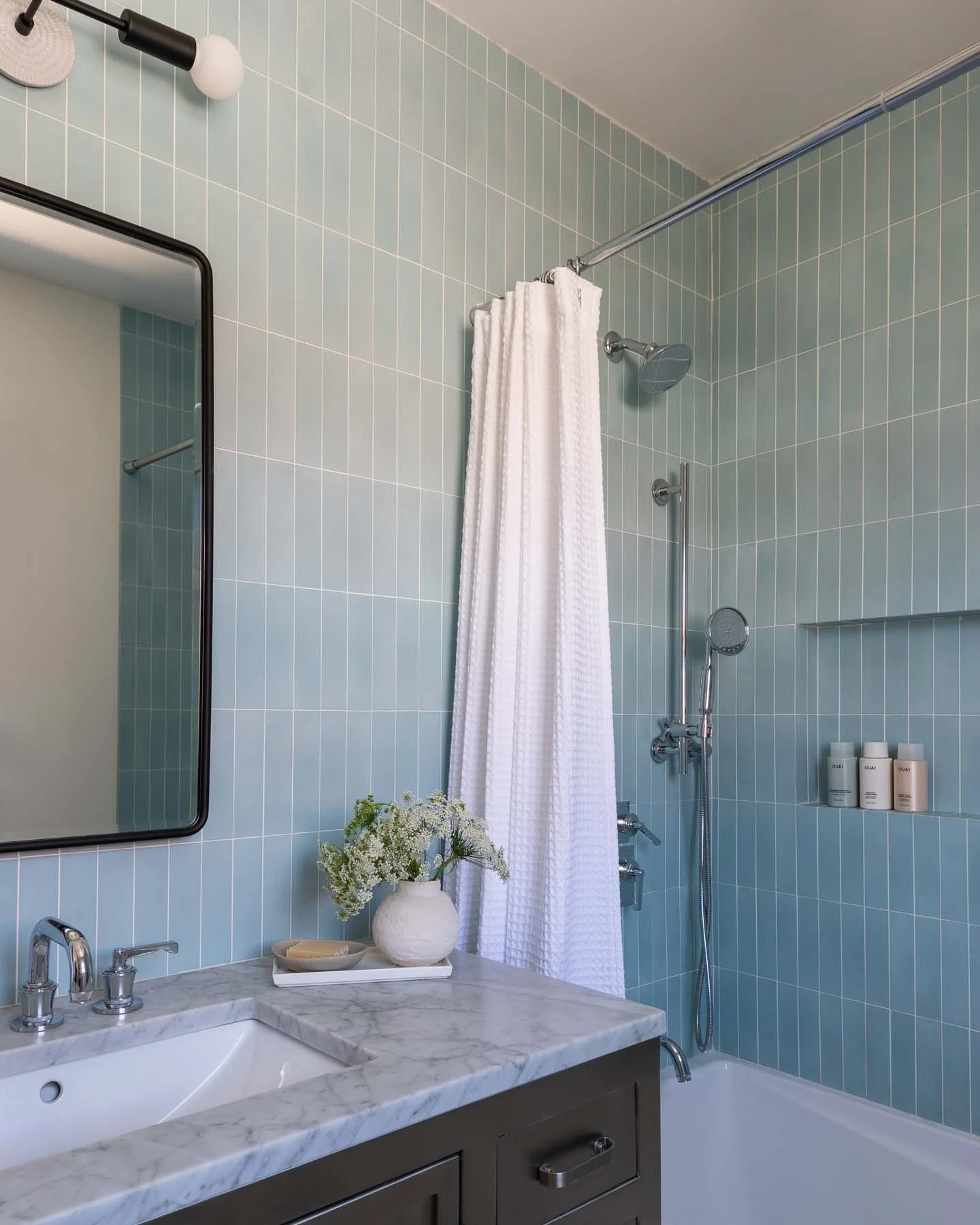 A bathroom with light blue tiled walls, a bathroom sink with a marble countertop, a mirror, a shower area with a white shower curtain, and toiletries on a built-in shelf.