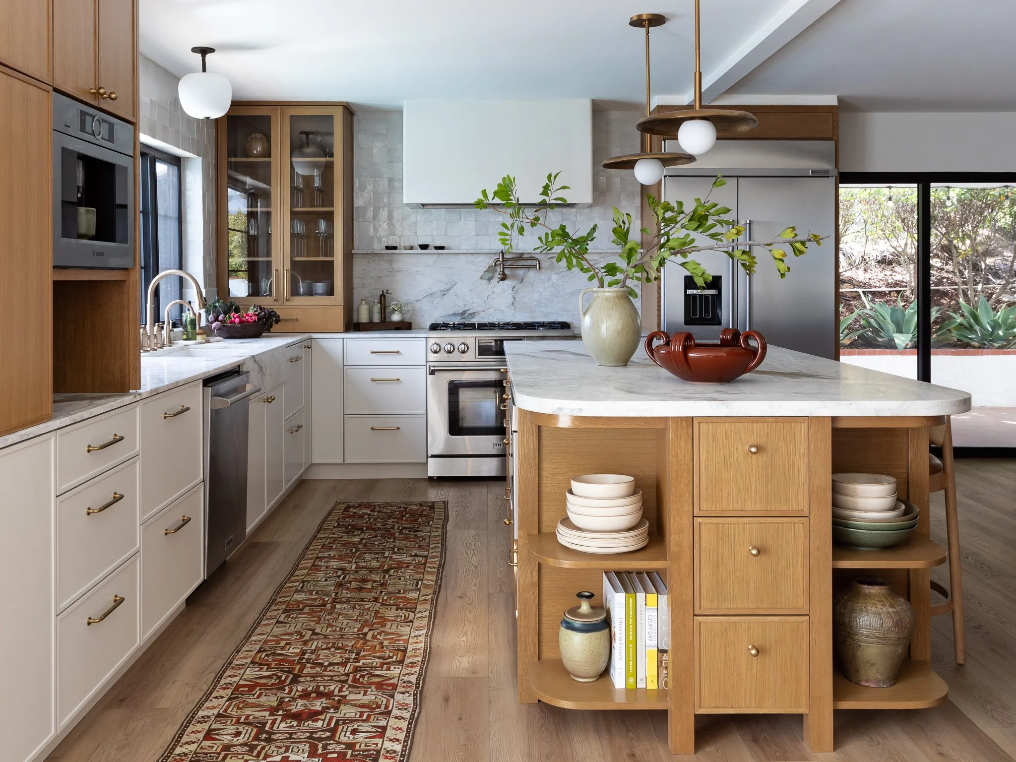 Modern kitchen with white cabinets, wooden accents, marble countertops, hanging light fixtures, and a decorative runner rug. There are plants, bowls, and books on a wooden shelf in the foreground.